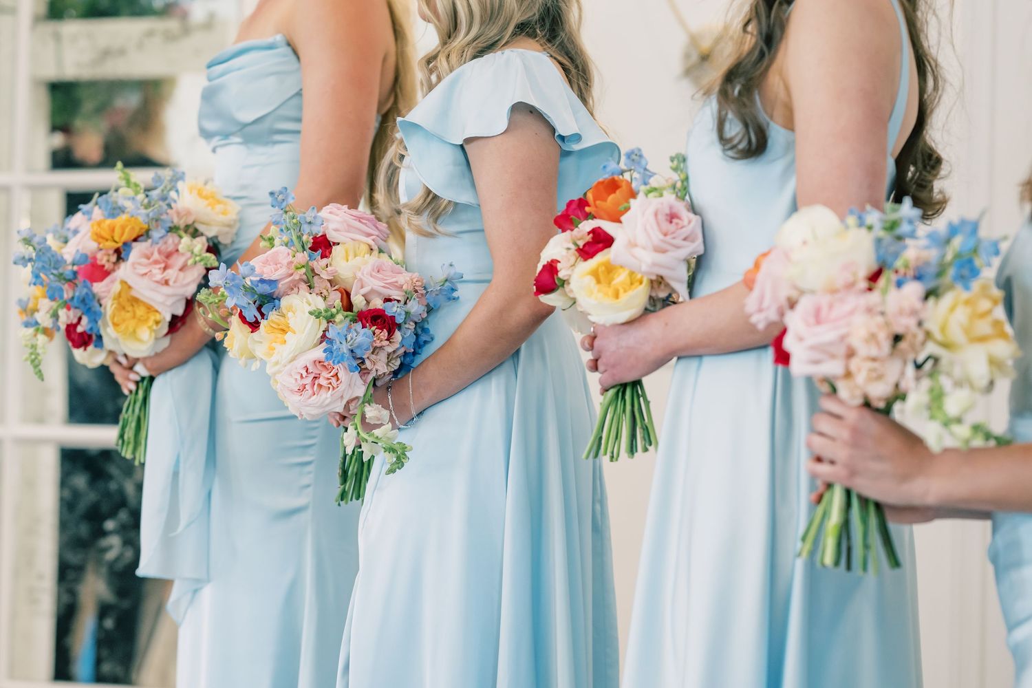 Bridesmaids in powder blue dresses holding lush colorful bouquets of roses, peonies, and wildflowers.