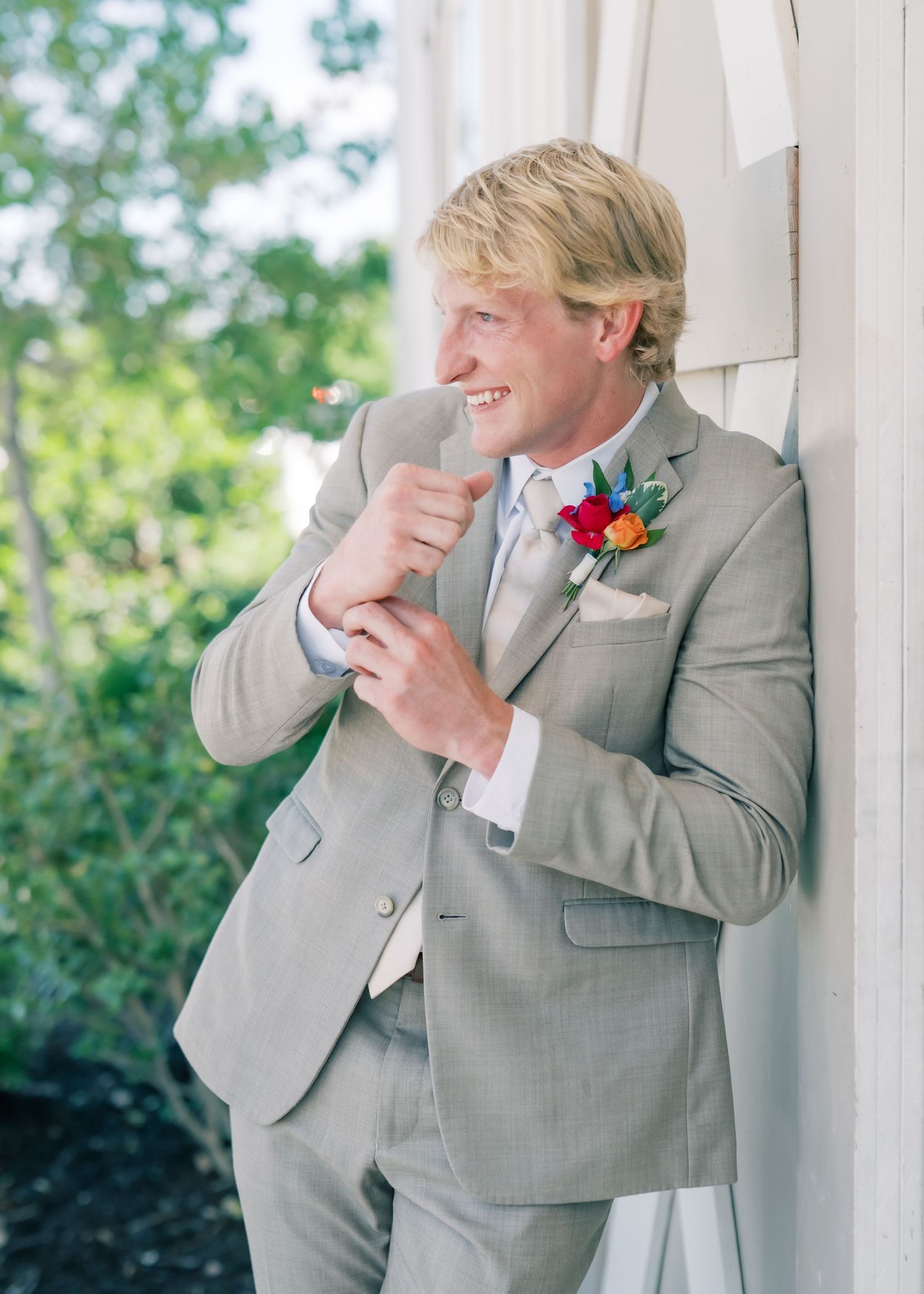 Smiling groom in a light gray suit adjusting his cufflinks, wearing a colorful boutonniere outdoors.