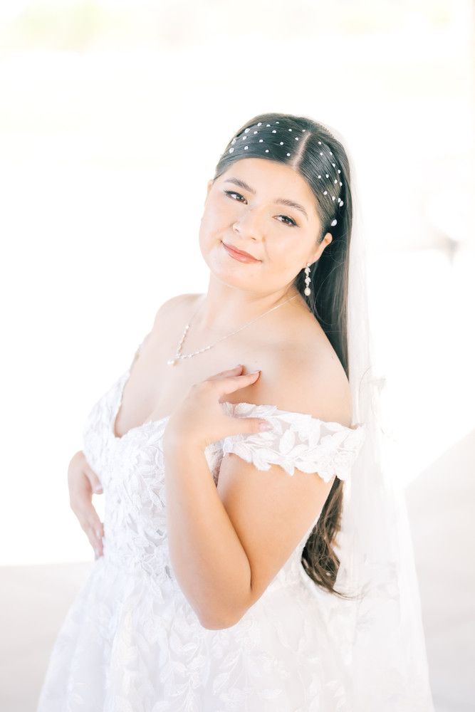 A bride in white lace dress poses against bright background wearing a pearl-adorned headpiece.