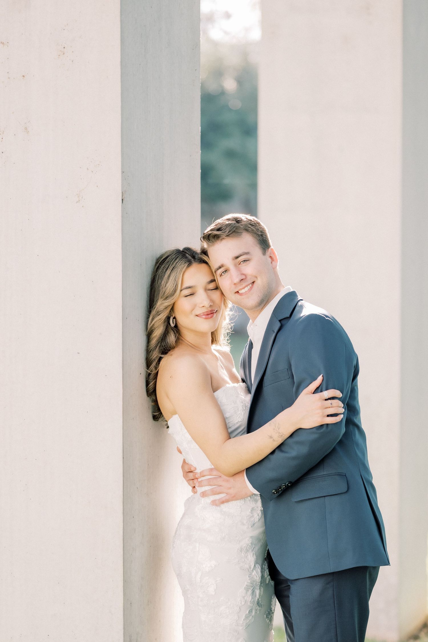 A happy couple in wedding attire embrace and smile while standing between white pillars in soft natural lighting.