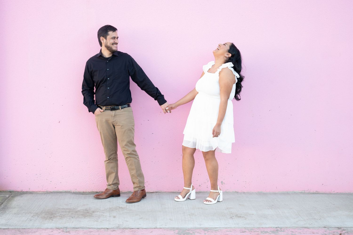 A couple poses playfully against a pink wall during a photo session.