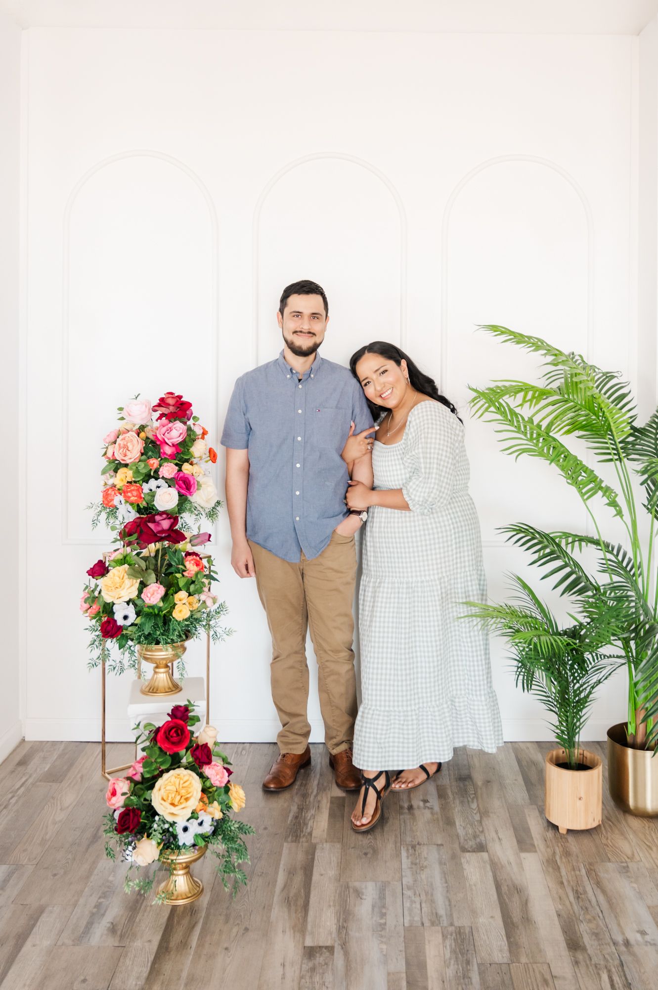 A couple poses in front of a white wall with floral arrangements and potted plants decorating the scene.