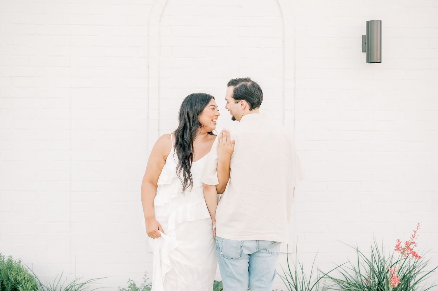 A couple in light colored outfits share intimate moments against a white wall in a romantic photo session.