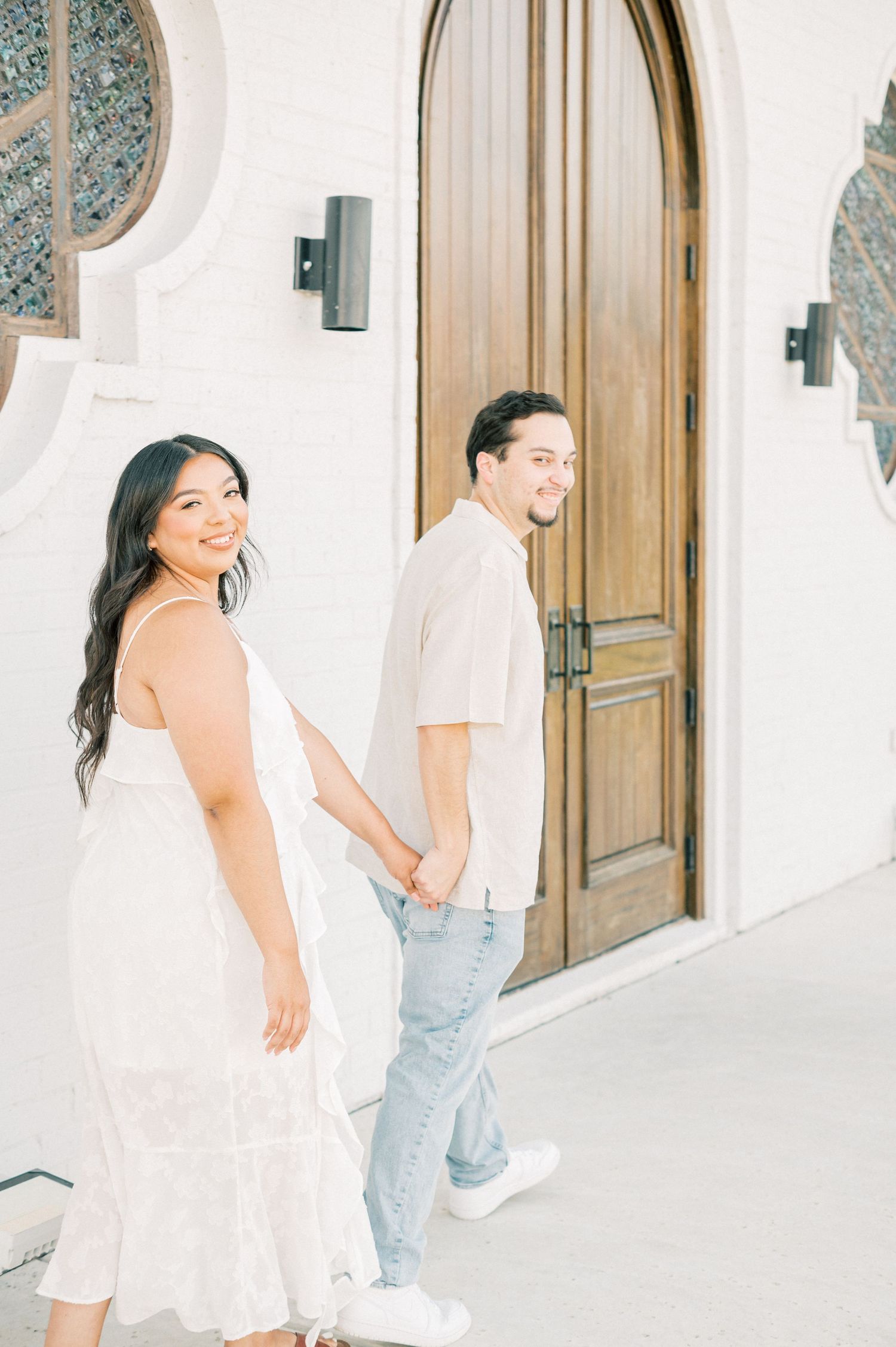 A couple shares an intimate moment against a white stucco wall with wooden doors in the background.