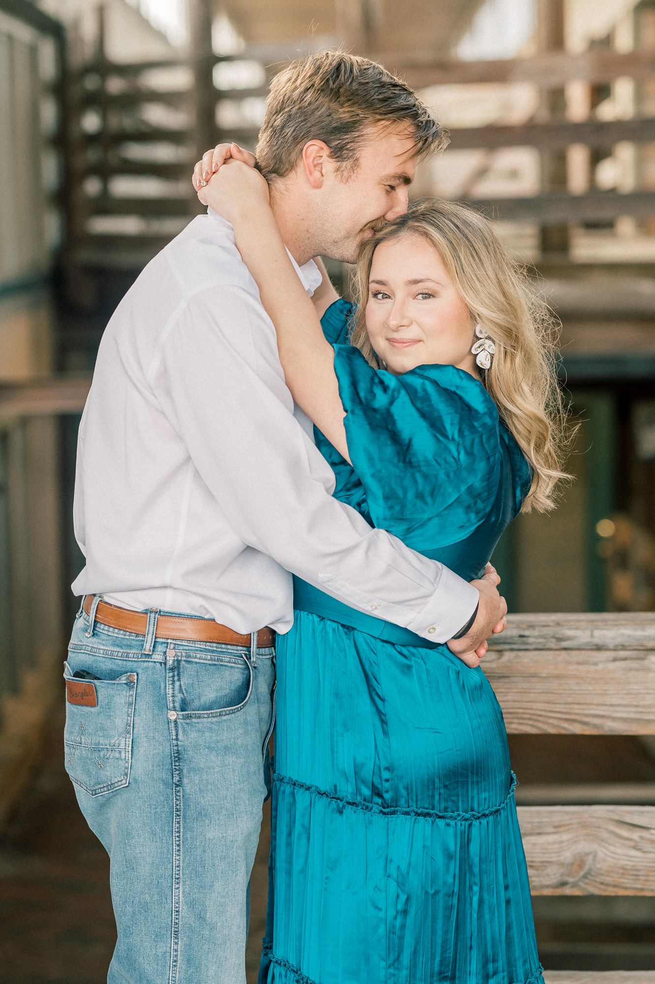 A romantic moment captured between a couple, with her in a teal dress and him in casual attire against a rustic backdrop.