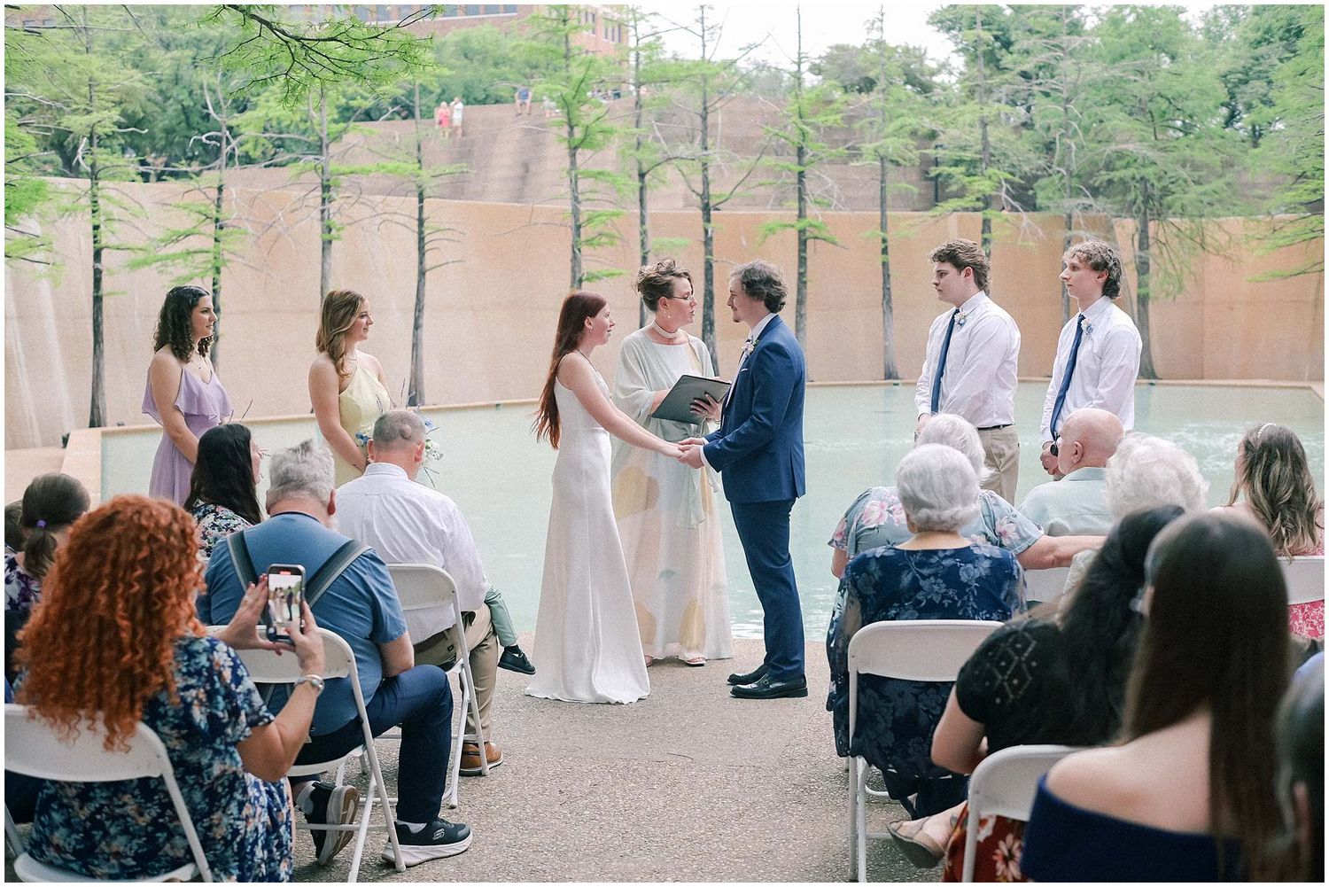 Guests seated in white folding chairs watch an outdoor wedding ceremony under tall pine trees.