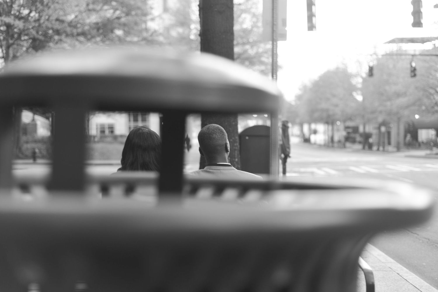 Black and white photo of two people sitting on a bench at an urban street corner, viewed from behind.