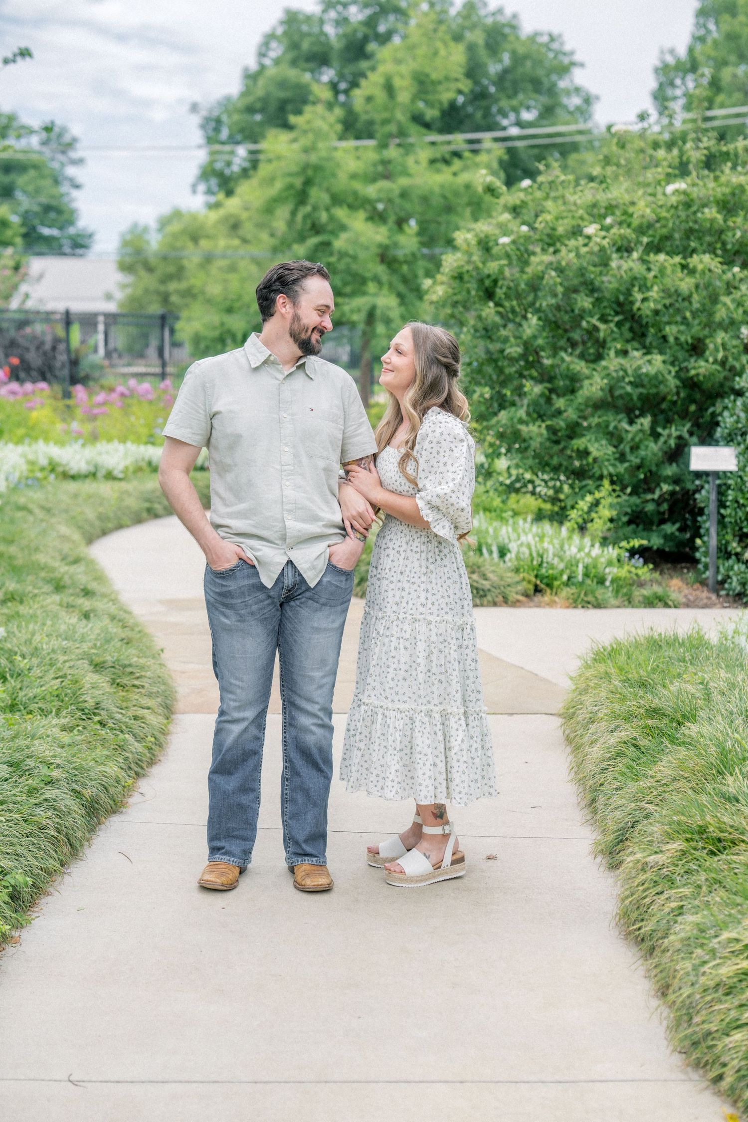 Couple smiling at each other on a garden path surrounded by lush greenery and colorful flowers.