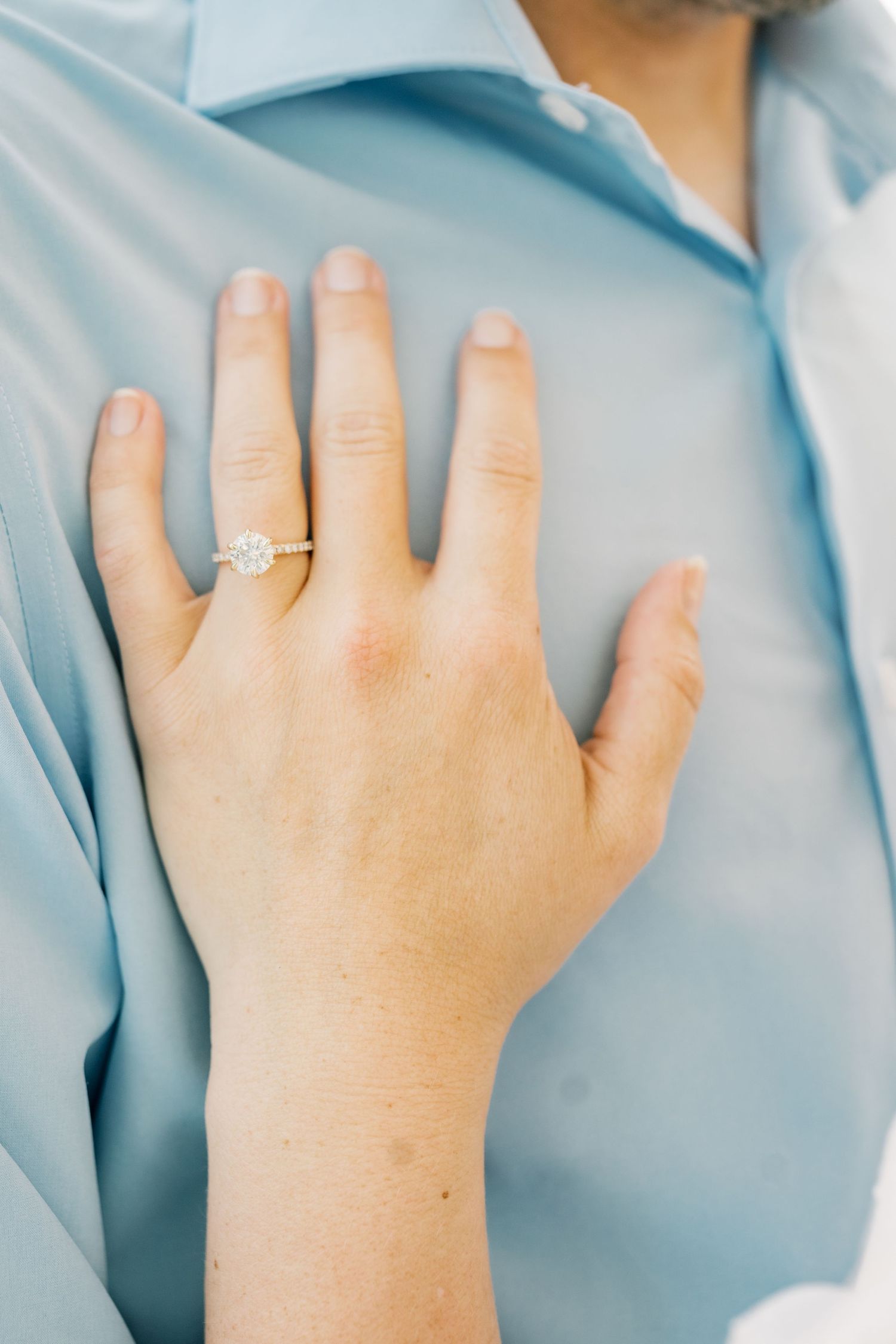 Close-up of a woman's hand displaying a diamond engagement ring resting on a man's light blue dress shirt.