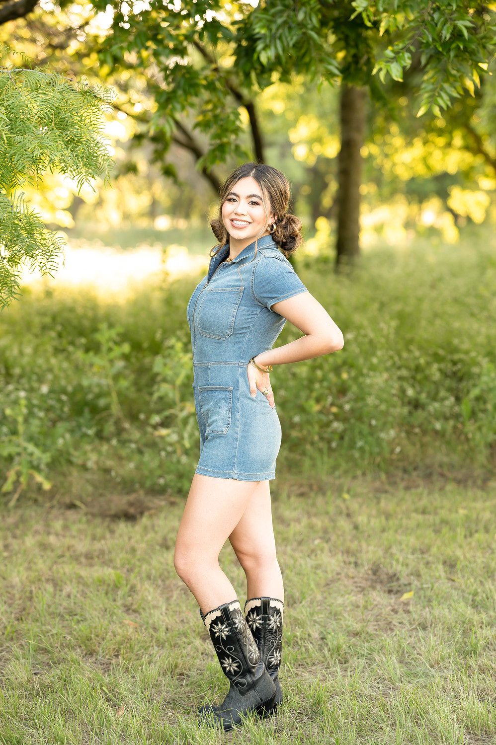Smiling young woman in denim mini dress and black cowboy boots posing outdoors in lush green park setting.