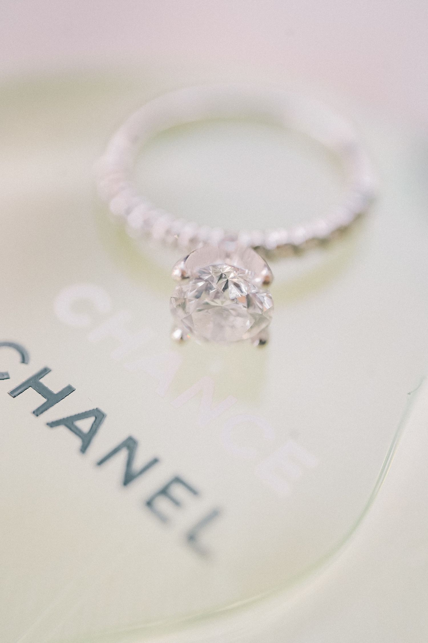 Elegant diamond solitaire engagement ring with pavé band resting on a Chanel box, showcasing brilliant cut stone.