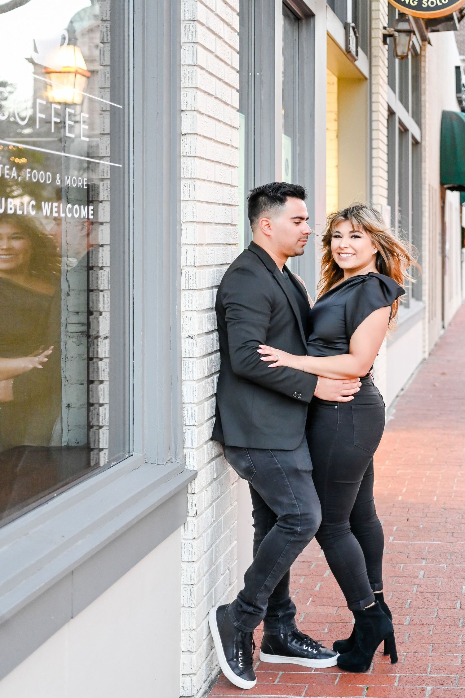 Couple in matching black outfits embracing against a brick wall outside a coffee shop on a city sidewalk.