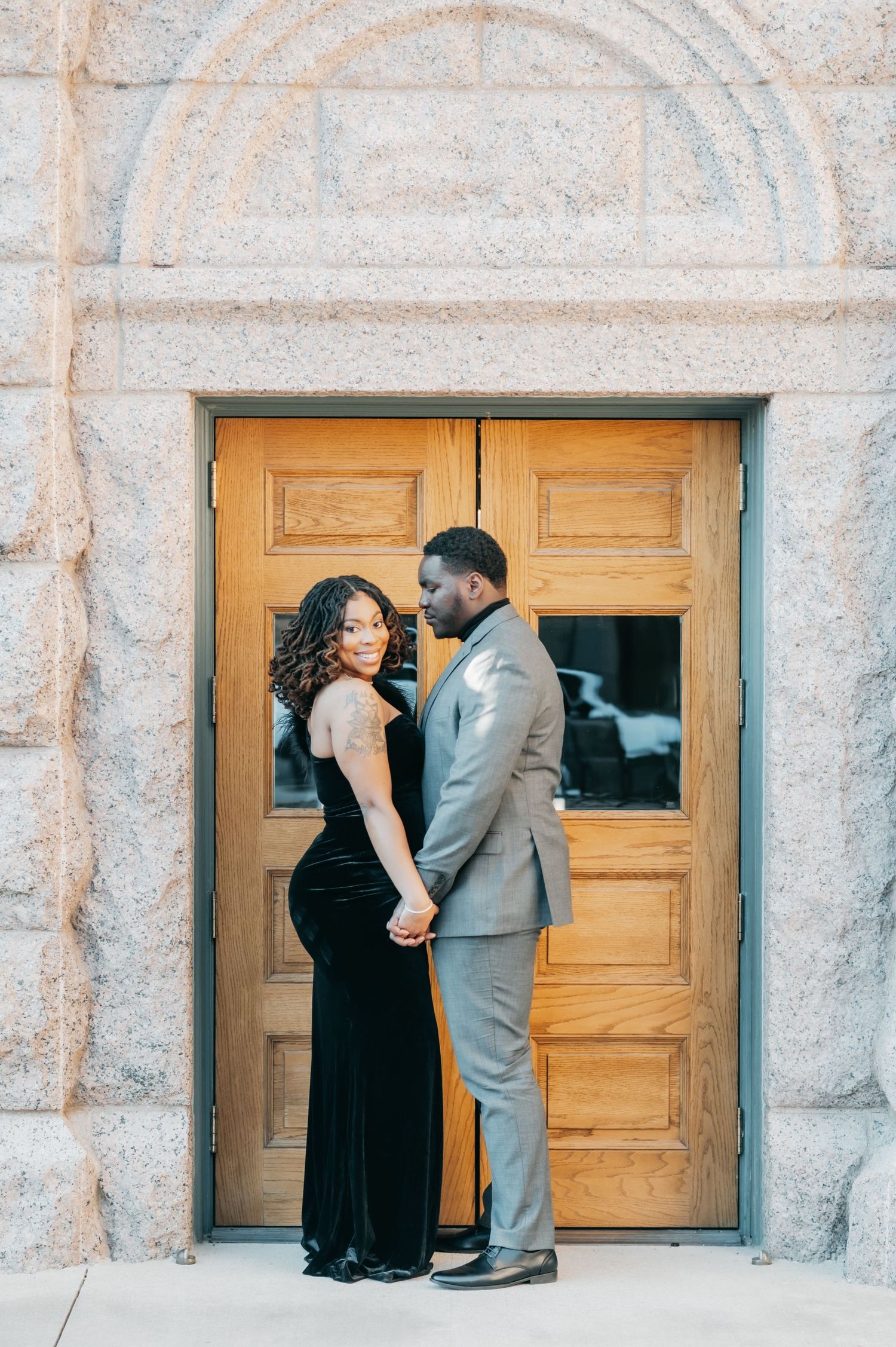 Romantic couple holding hands and gazing at each other before wooden doors in a stone archway.