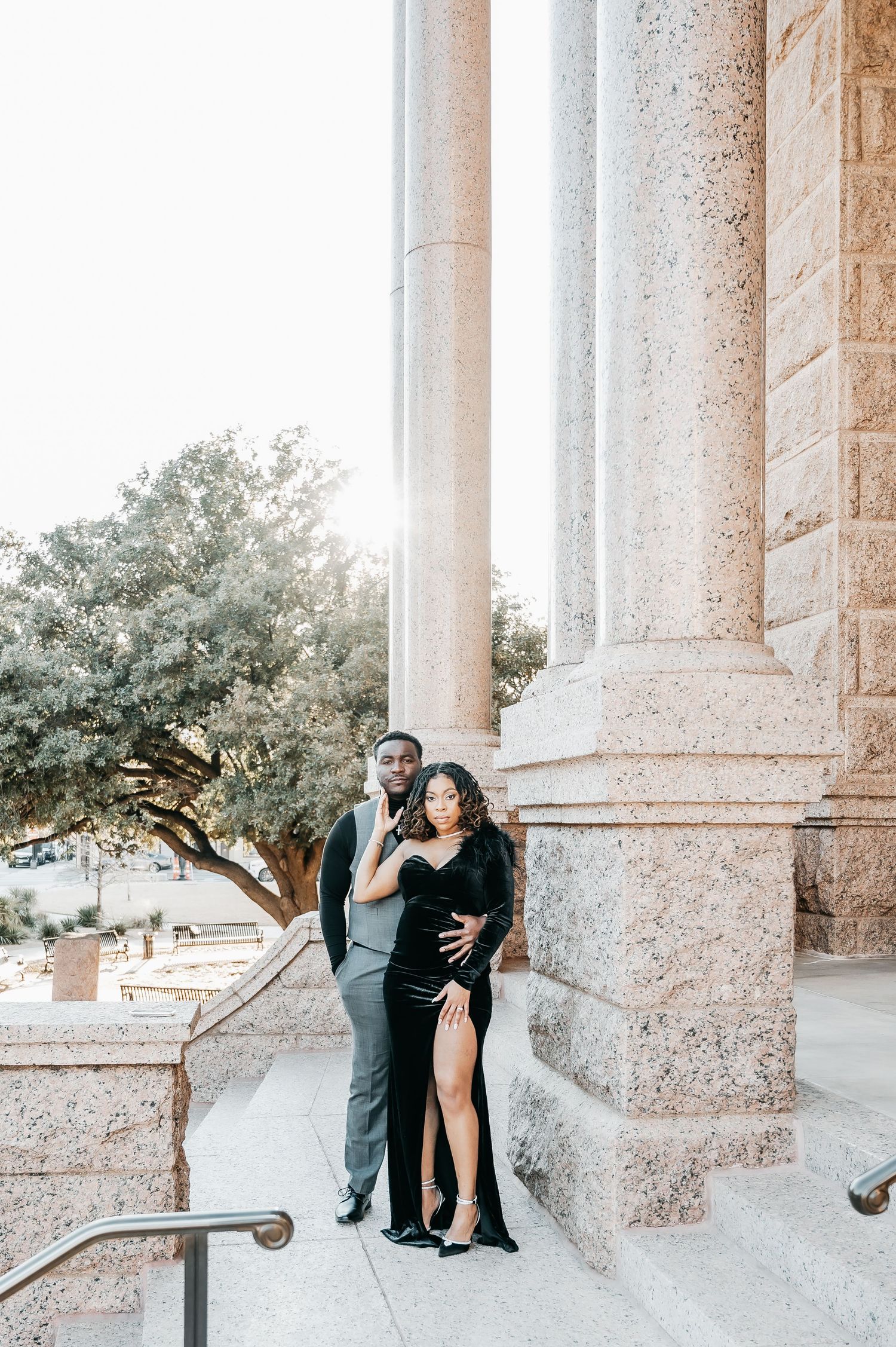 Elegant couple posing by stone columns in black formal attire during a golden hour engagement photo session.