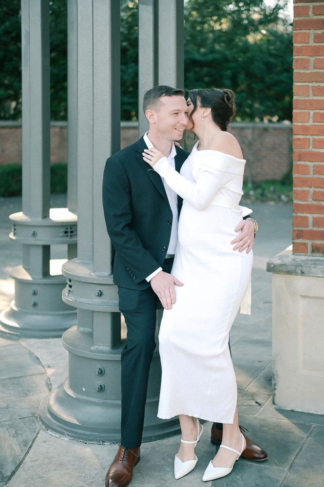 Bride in elegant off-shoulder white gown kissing groom in dark suit beside stone columns outdoors.