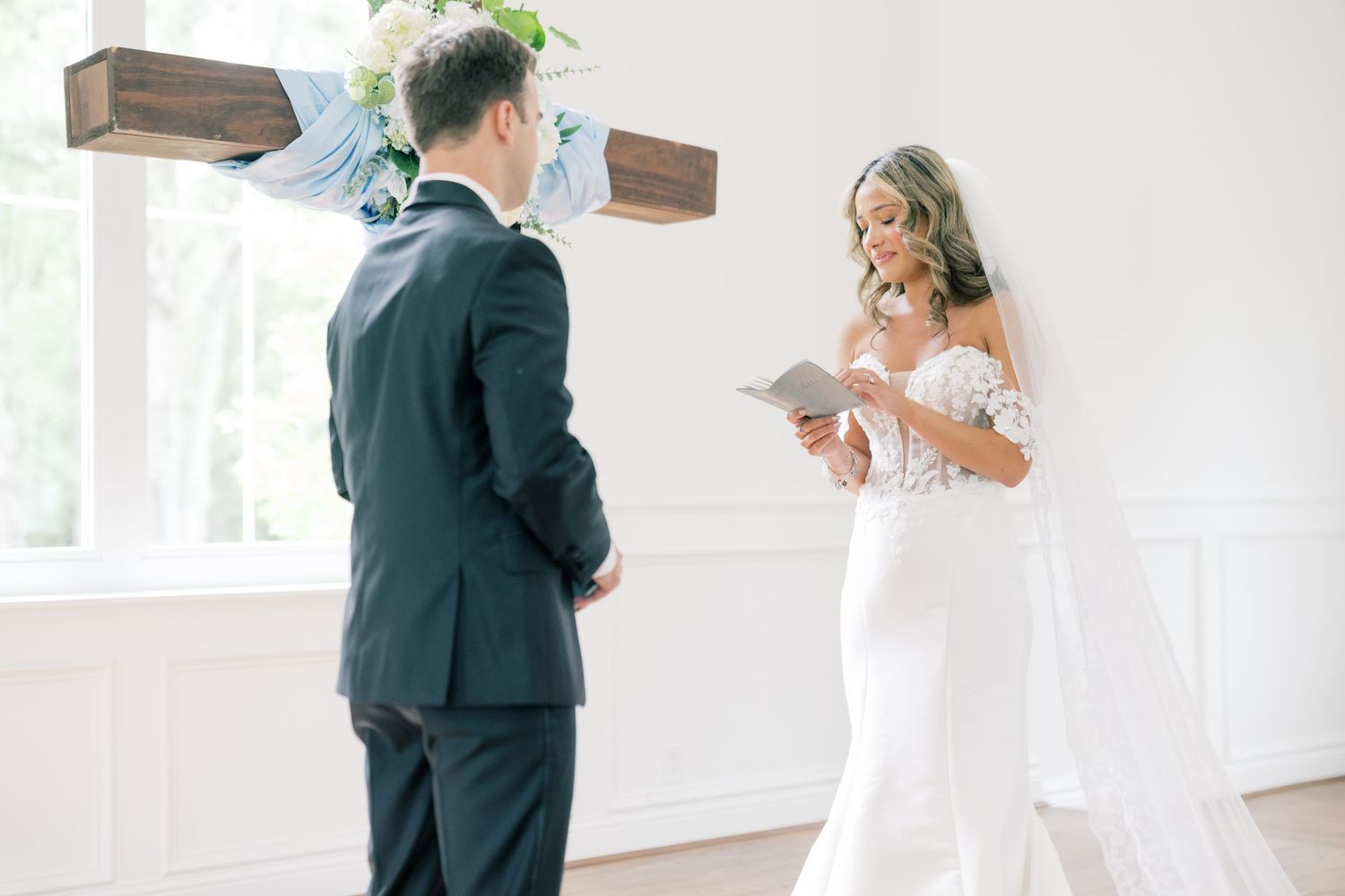 Bride in lace wedding dress reading vows to groom at altar with wooden cross.