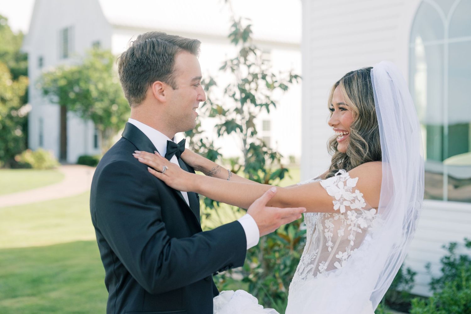 Groom reacts with joy seeing bride in lace wedding dress during outdoor first look moment.
