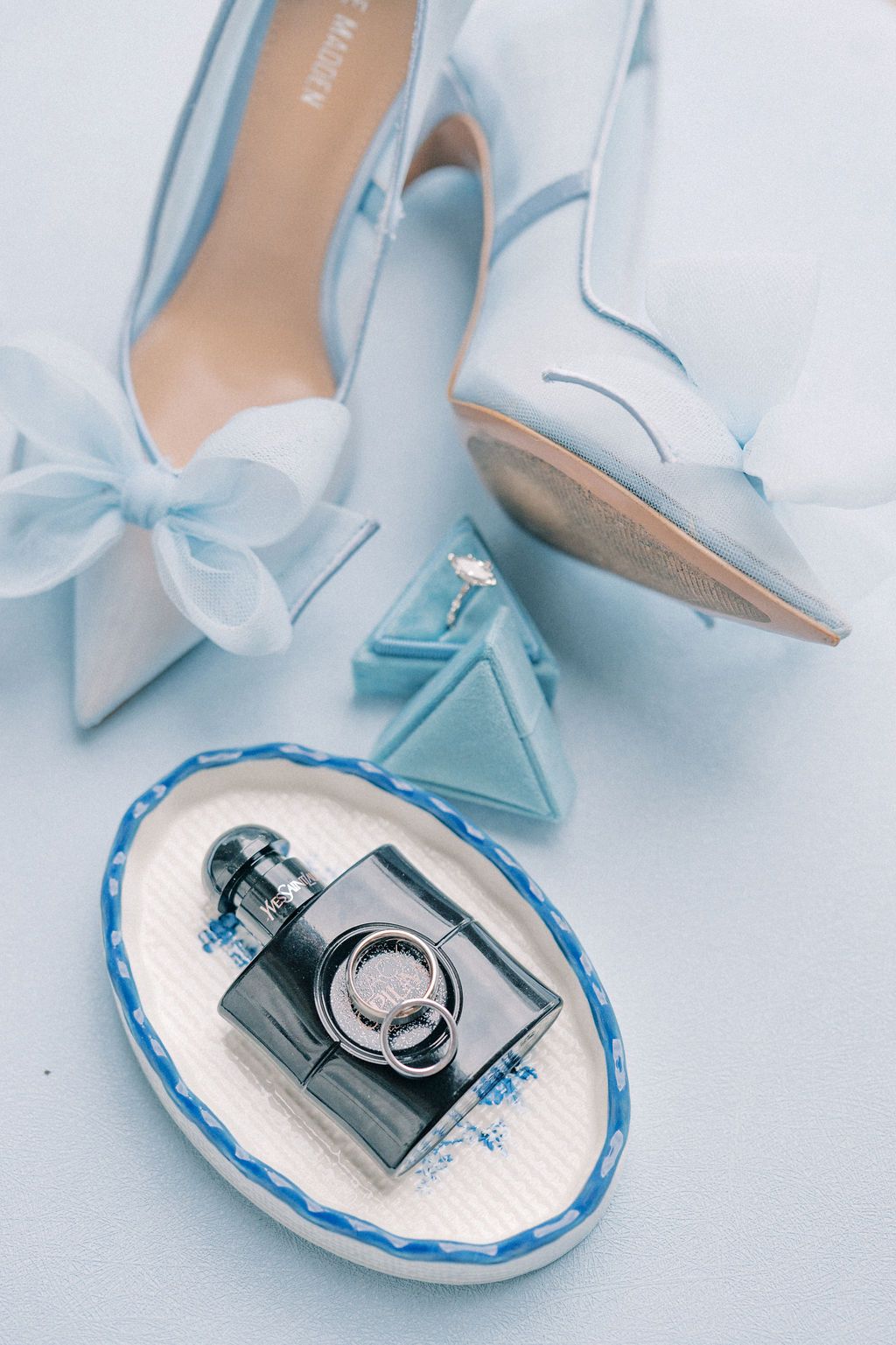 Light blue bridal accessories flat lay with perfume bottle, wedding rings on oval dish, and bow-detail heels.