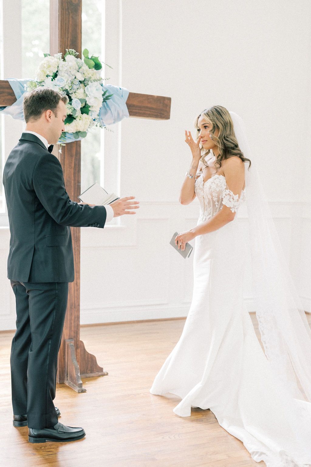Bride wiping tears as groom reads wedding vows at altar with floral cross decoration in elegant white chapel.