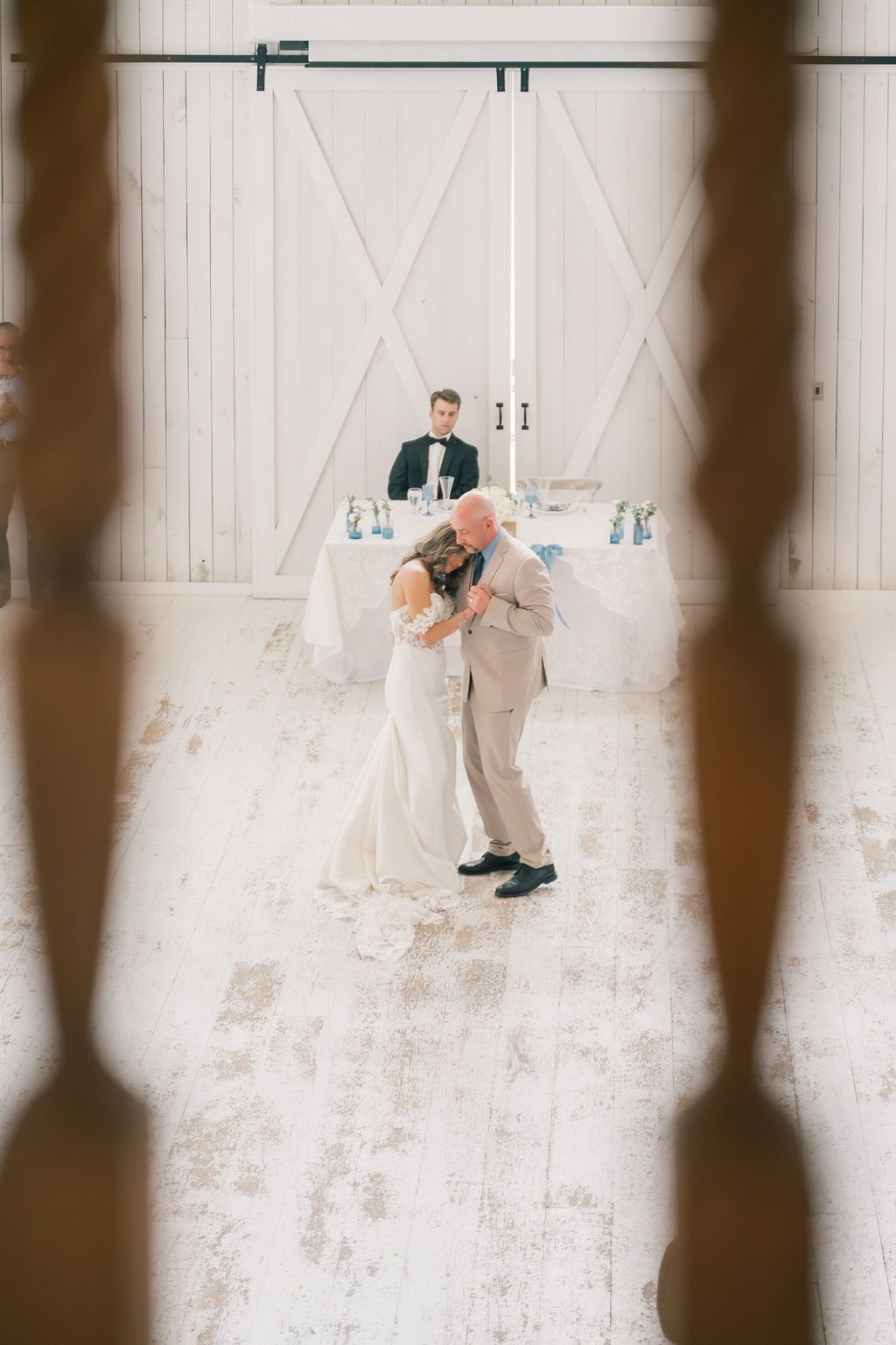 Bride and father share emotional first dance in a white barn venue while groom watches from sweetheart table.