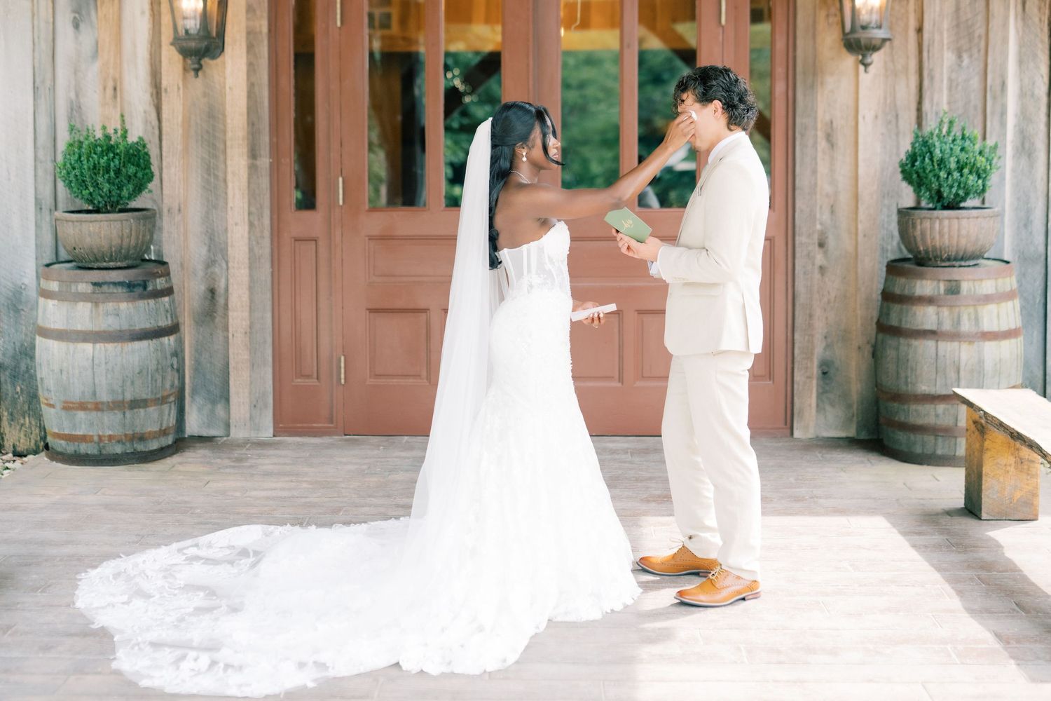 Bride in white gown with long veil stands with groom in tan suit outside rustic barn wedding venue with wine barrels.