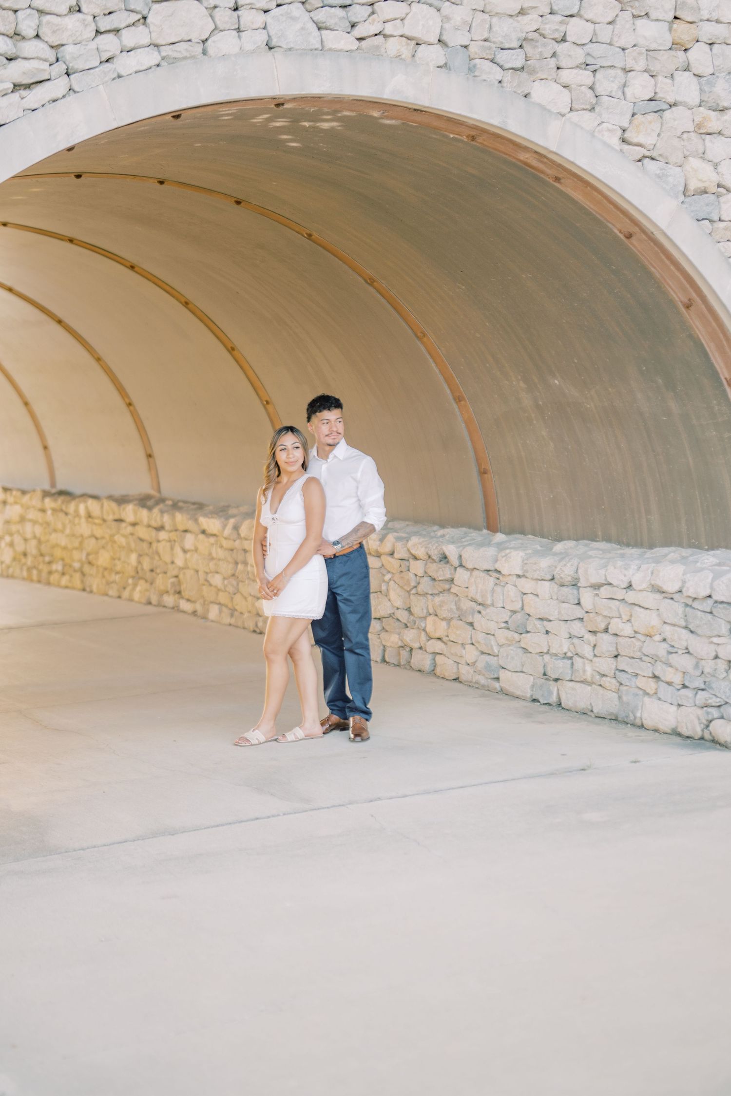 Couple in white outfits posing romantically under a stone arch tunnel during an engagement photo session.