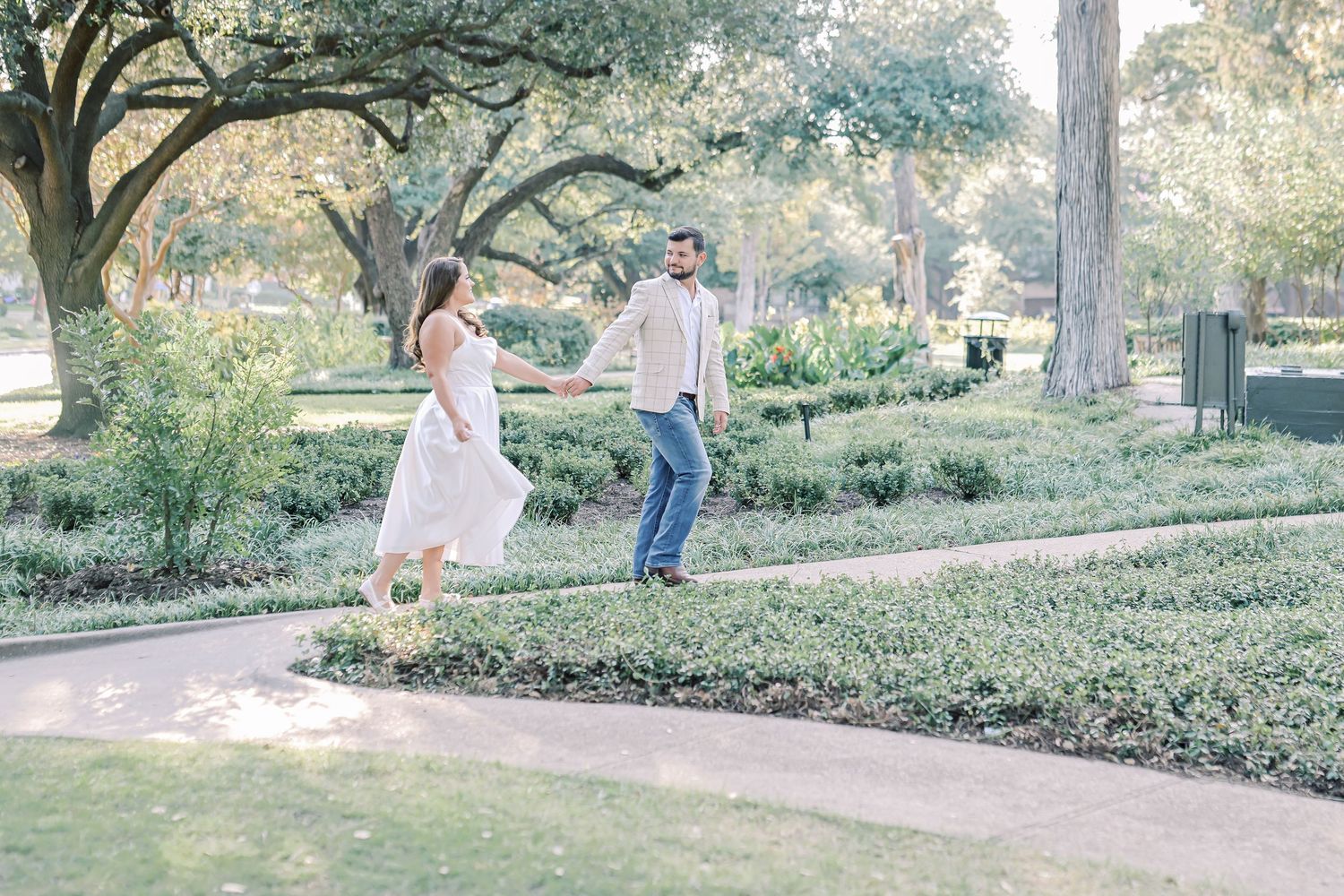 Couple holding hands and walking along a curved path through a lush, tree-filled park, the woman in a flowing white dress.