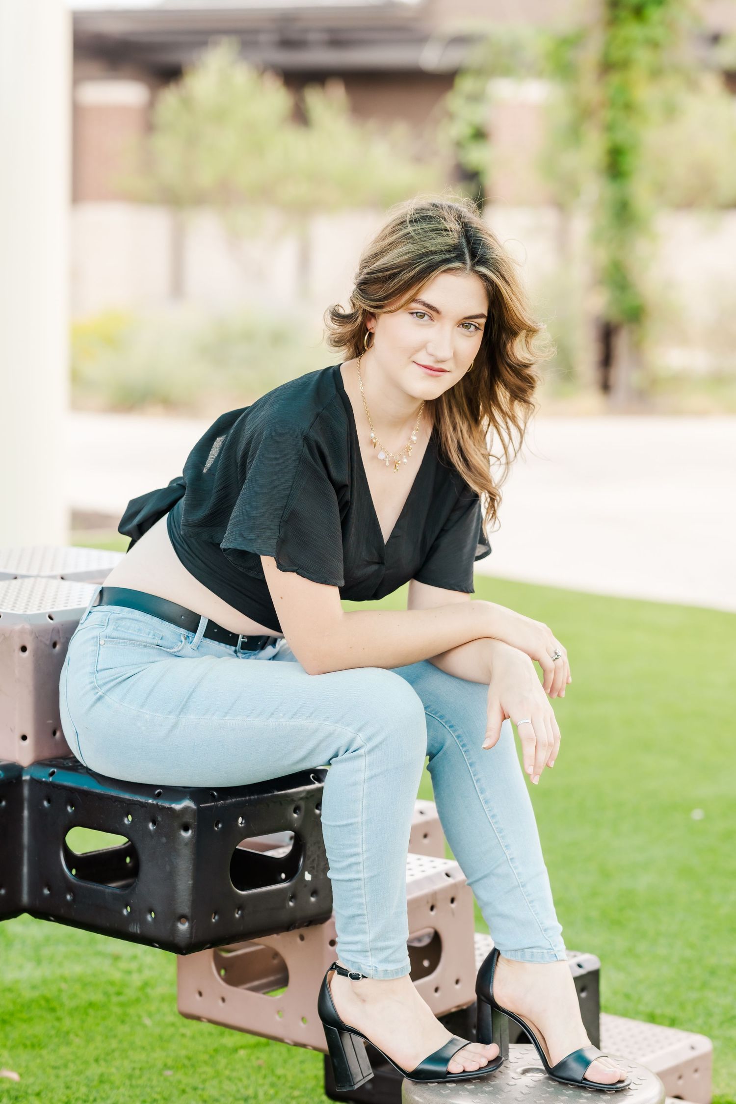 Young woman in black crop top and light jeans sitting on a bench outdoors, smiling confidently at the camera.