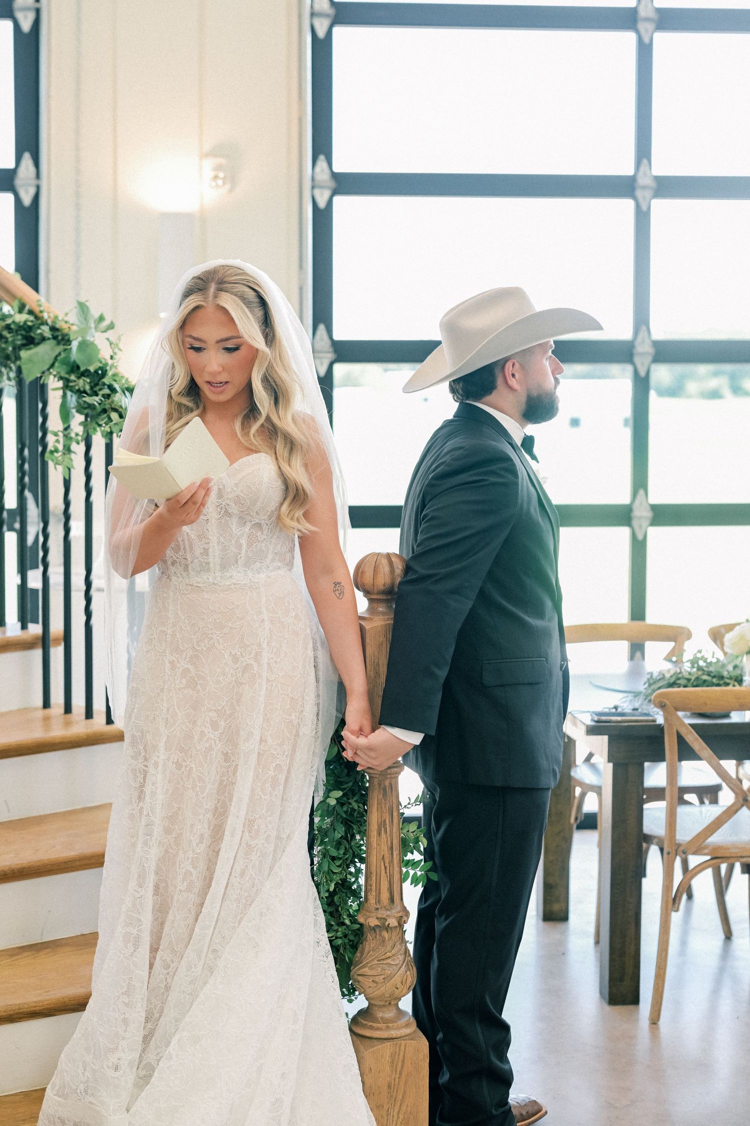Bride in lace wedding gown reads vows while groom in cowboy hat holds her hand at staircase.