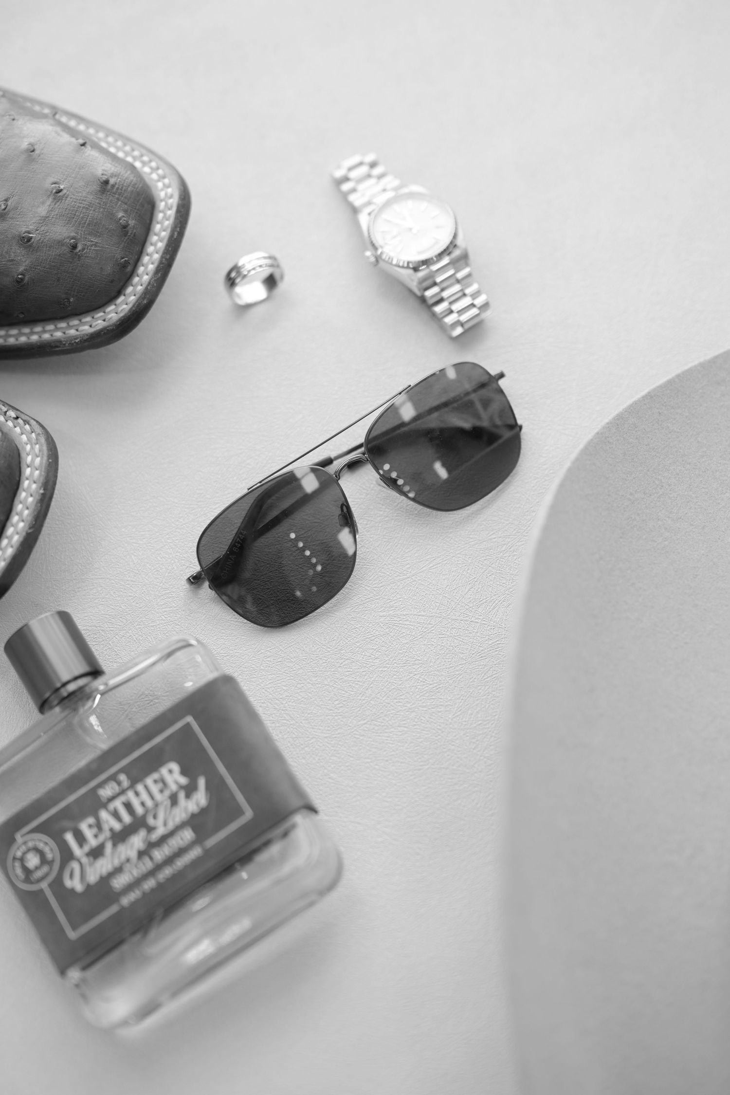 Black and white flat lay of groom accessories including sunglasses, watch, cologne, and ring.