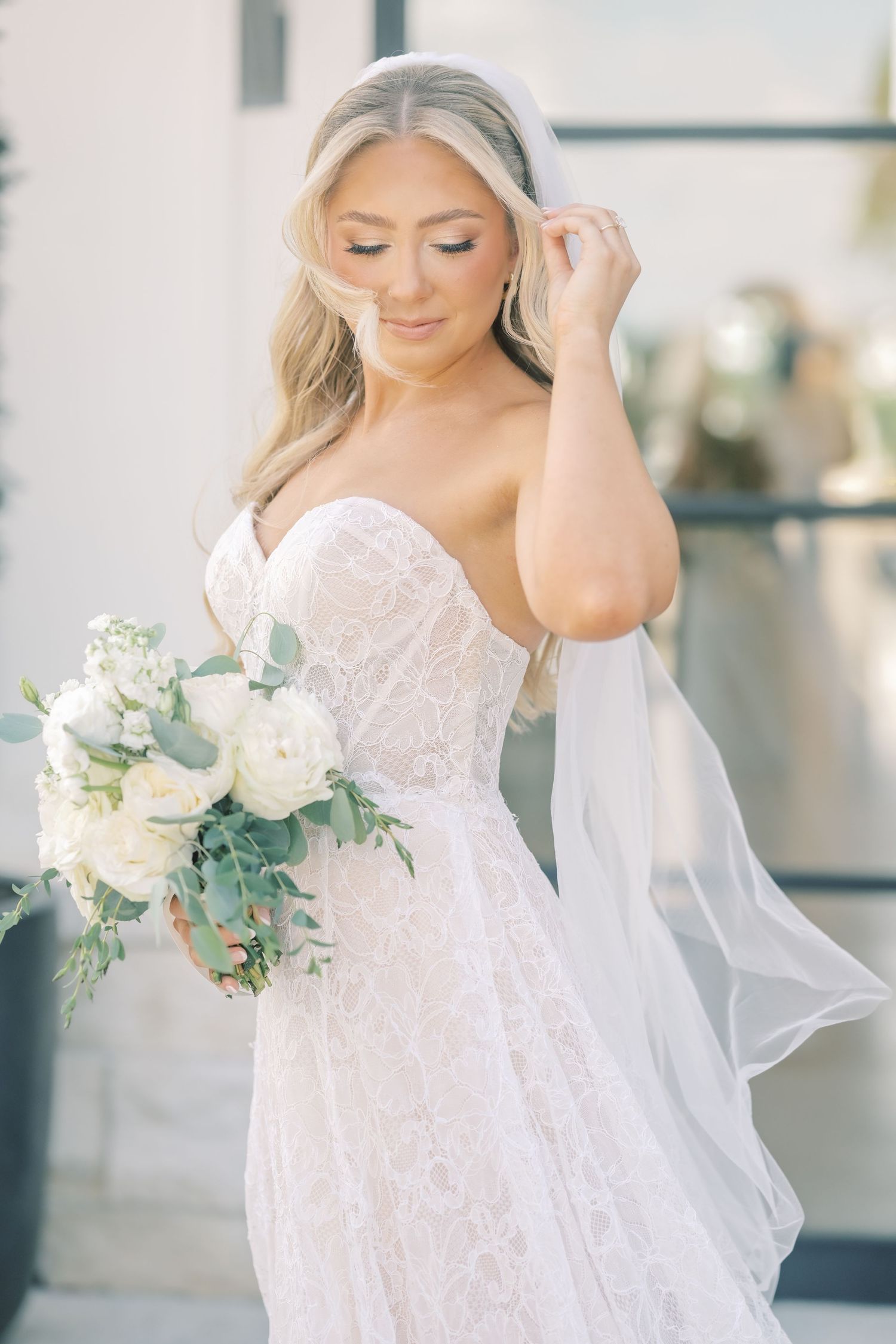 Elegant bride in strapless lace gown adjusts veil while holding white floral bouquet.