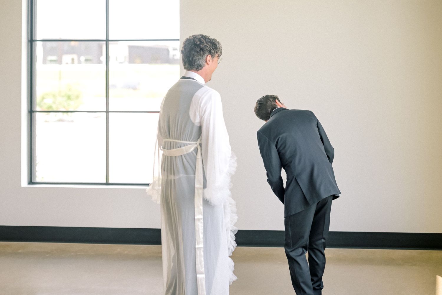 A priest in white robes stands with a suited man bowing near a bright window in a minimalist room.