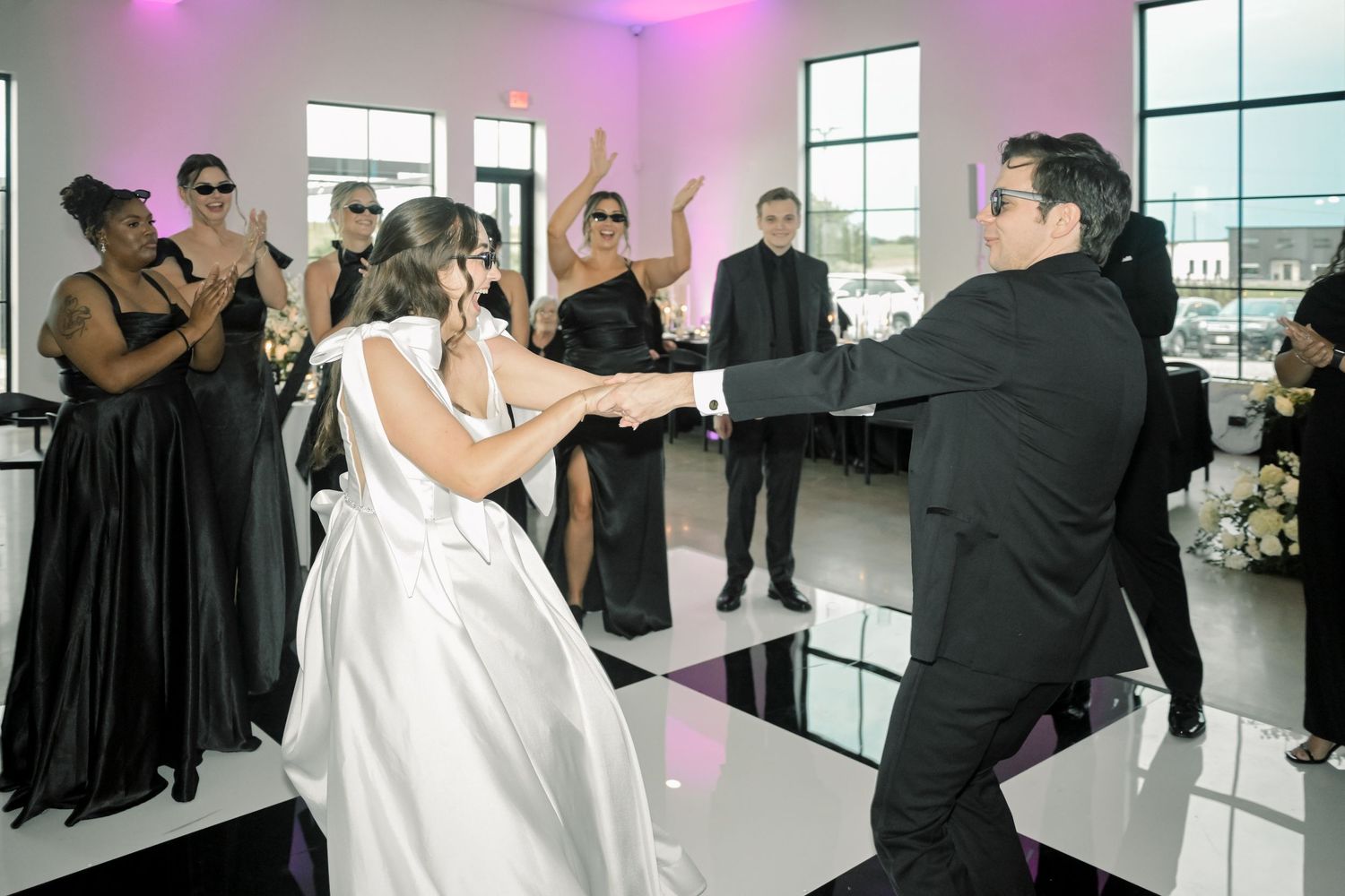 Bride and groom sharing a joyful first dance surrounded by cheering guests in black at their reception.