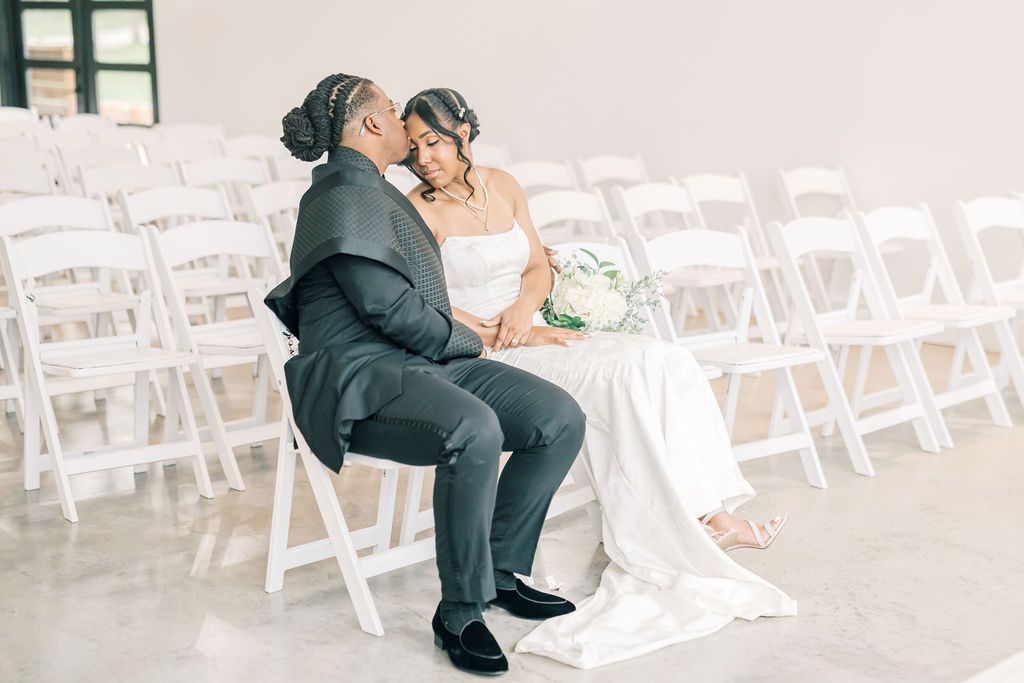 A bride and groom share an intimate moment seated on white folding chairs in an elegant wedding ceremony venue.