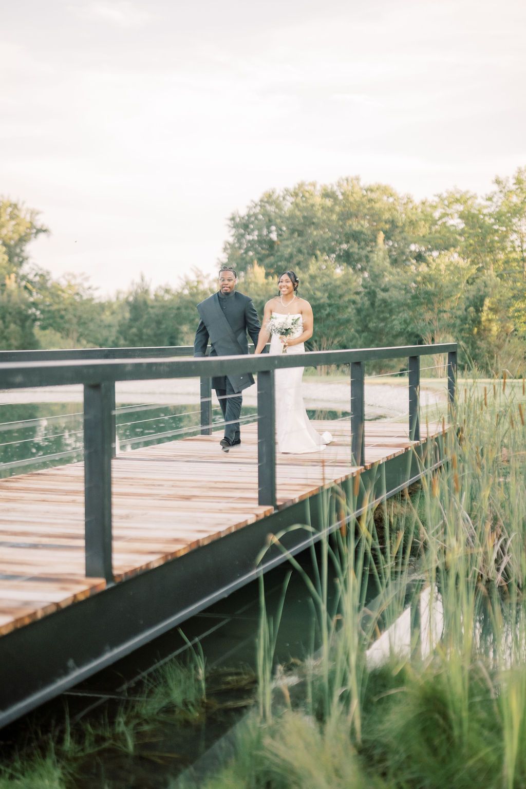 Bride and groom walking hand in hand on a rustic wooden bridge surrounded by lush greenery and a serene pond.