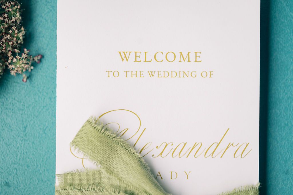 Gold foil wedding welcome sign for Alexandra on teal background with green ribbon and baby's breath.