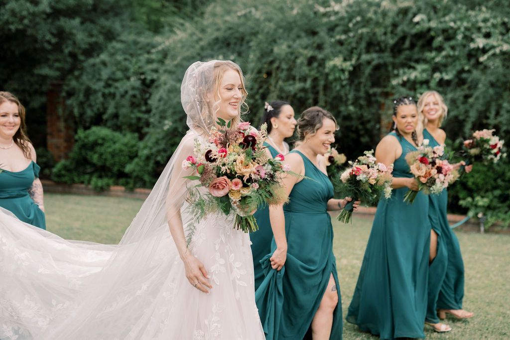 Bride in white gown with veil walks alongside bridesmaids in teal dresses holding colorful bouquets outdoors.
