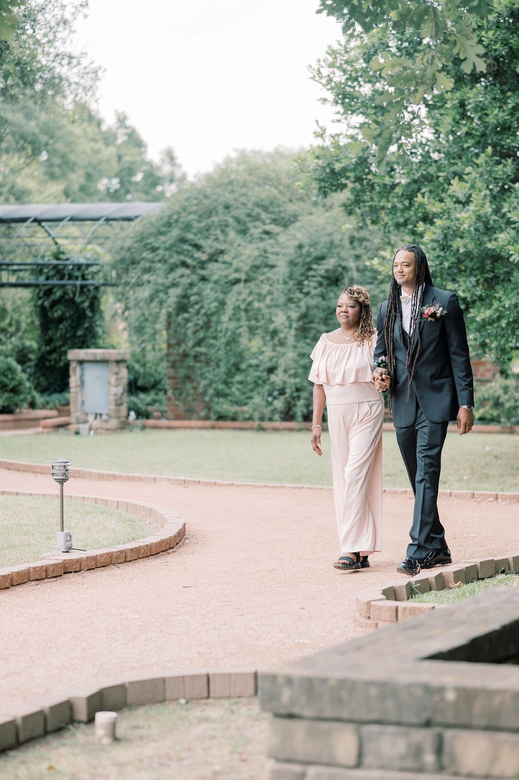 Couple walks hand-in-hand along a brick garden path, woman in white jumpsuit and man in dark suit, surrounded by lush greenery.