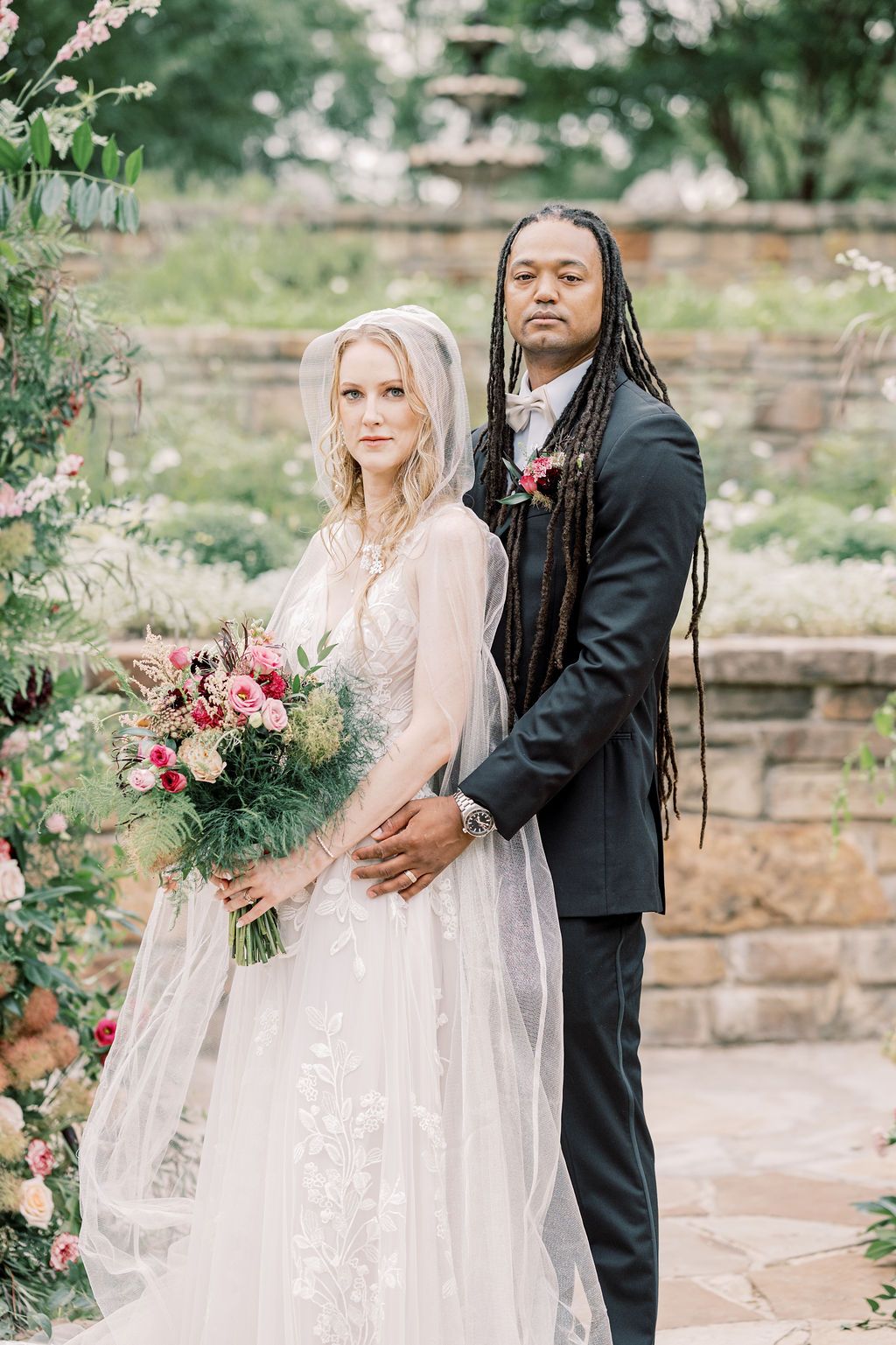 Interracial couple posing in garden wedding, bride in lace gown with veil holding bouquet, groom in dark suit with dreadlocks.