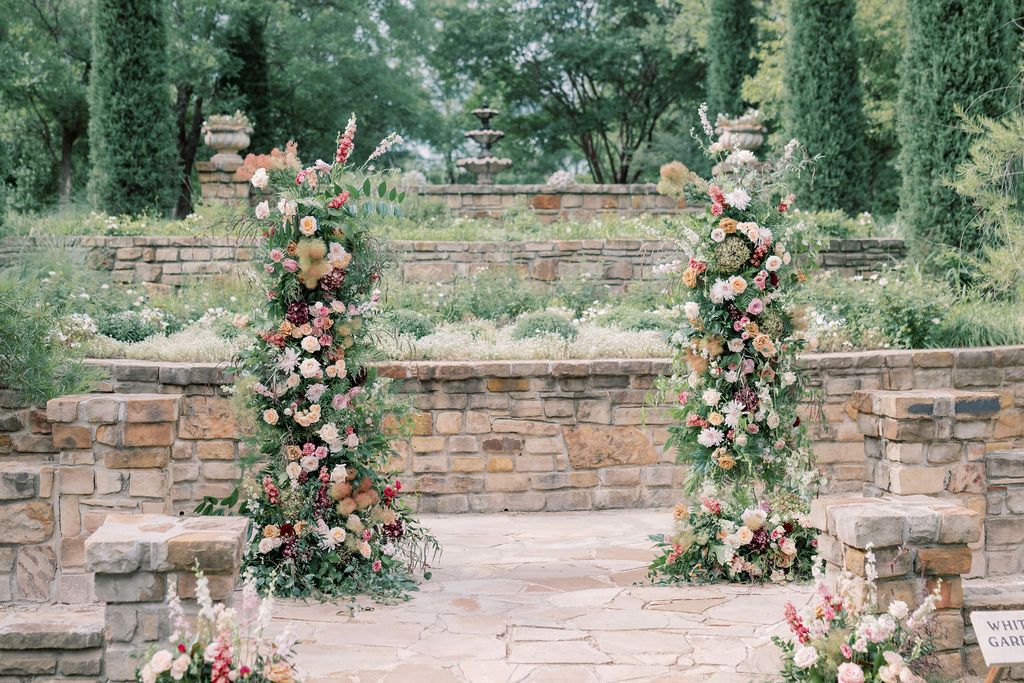 Two floral columns with pink and green blooms flank a stone wedding ceremony arch in a lush garden setting.