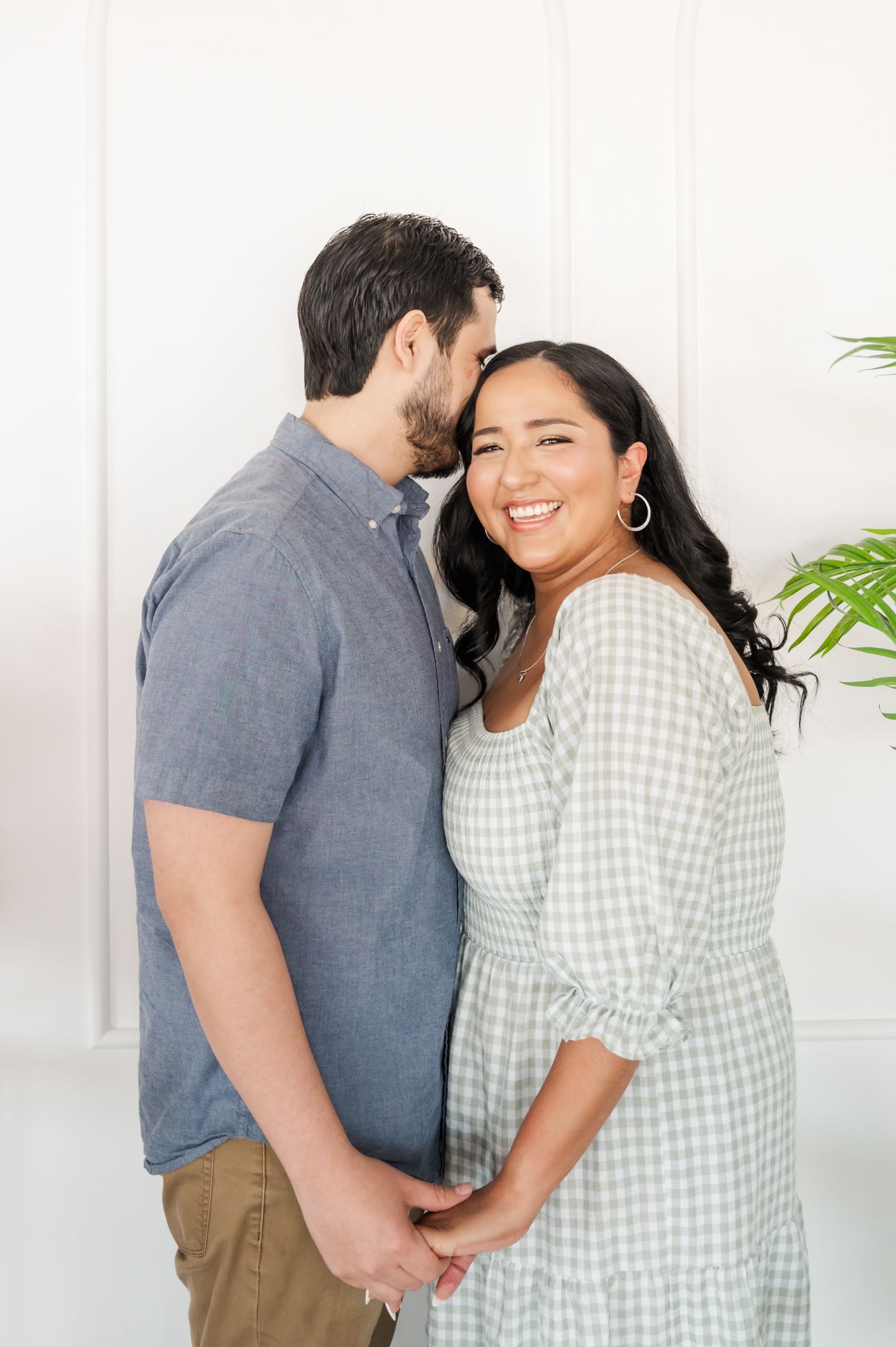A romantic couple embraces in front of a white wall with green plants.