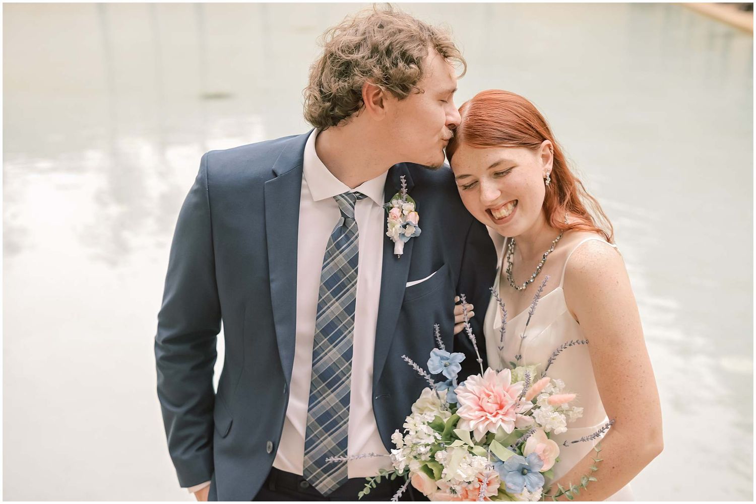 Intimate wedding portrait against a light colored wall showcasing a romantic embrace and floral bouquet.