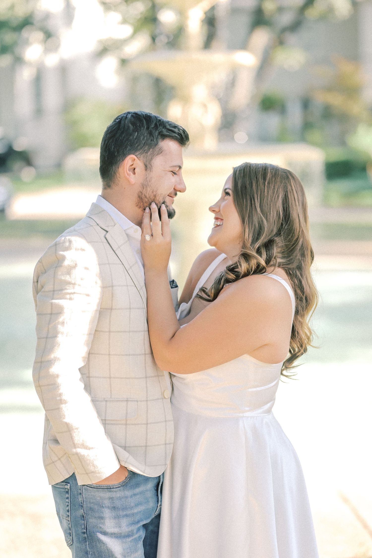 Smiling couple in an outdoor engagement photo, the woman showing her ring as she touches the man's face near a fountain in soft sunlight.
