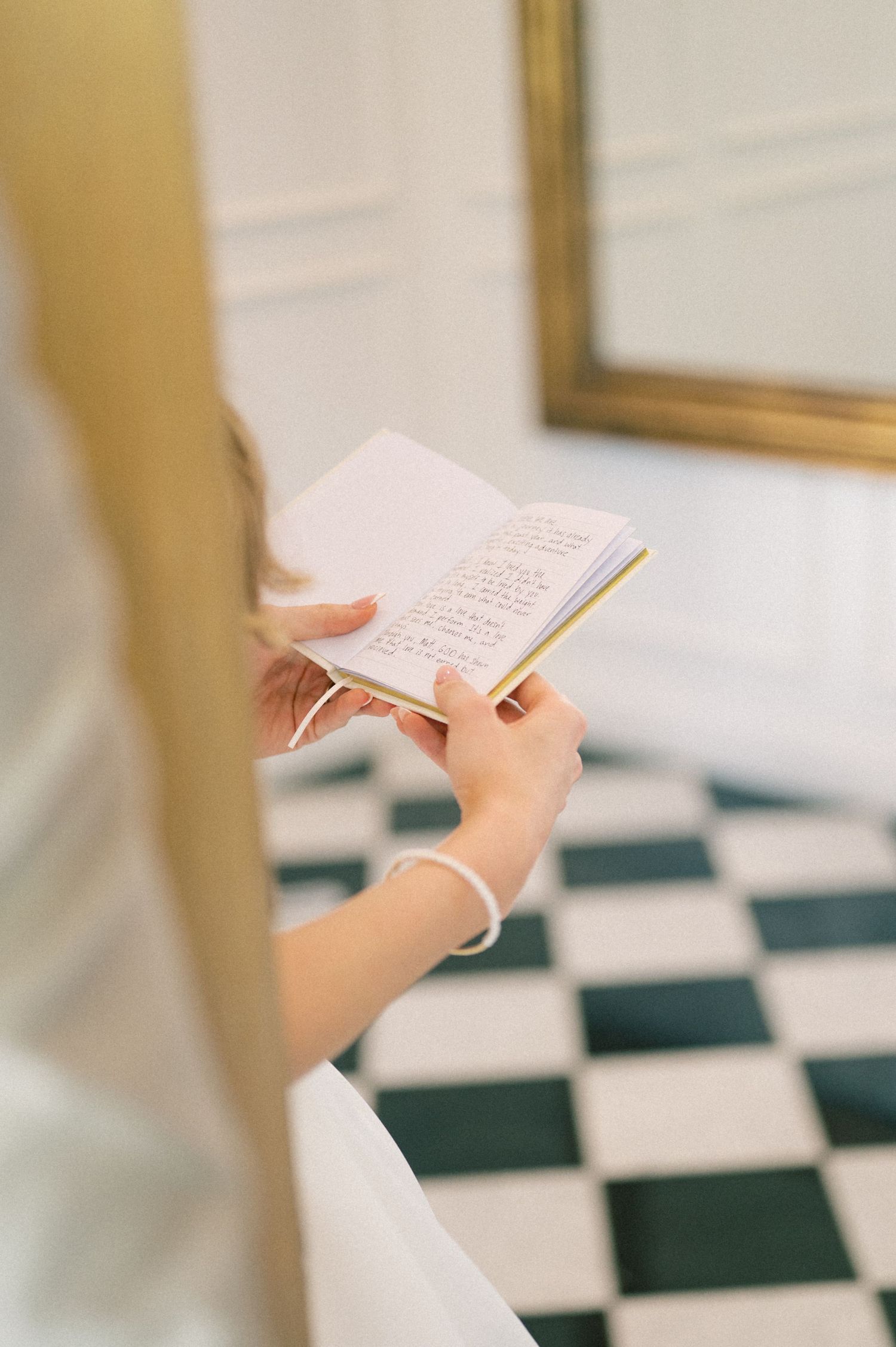 Bride in white dress reads handwritten vows from small notebook on checkered black and white floor.