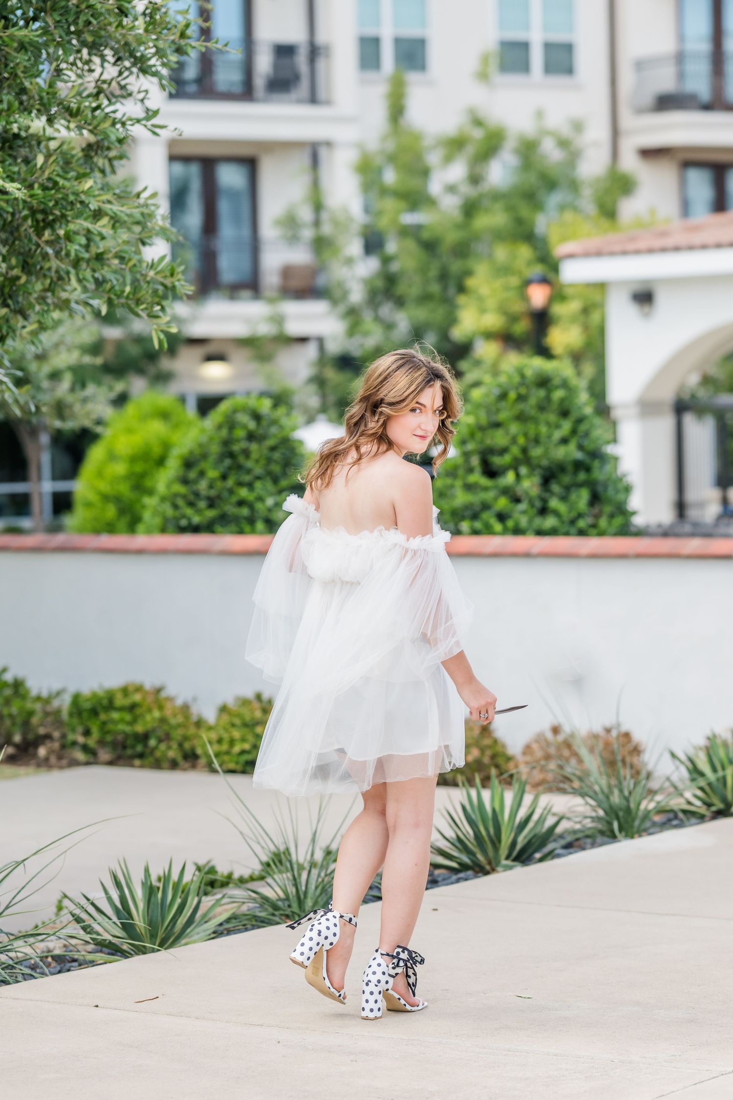 Woman in white off-shoulder tulle dress and polka dot heels posing outdoors near manicured gardens and elegant building.