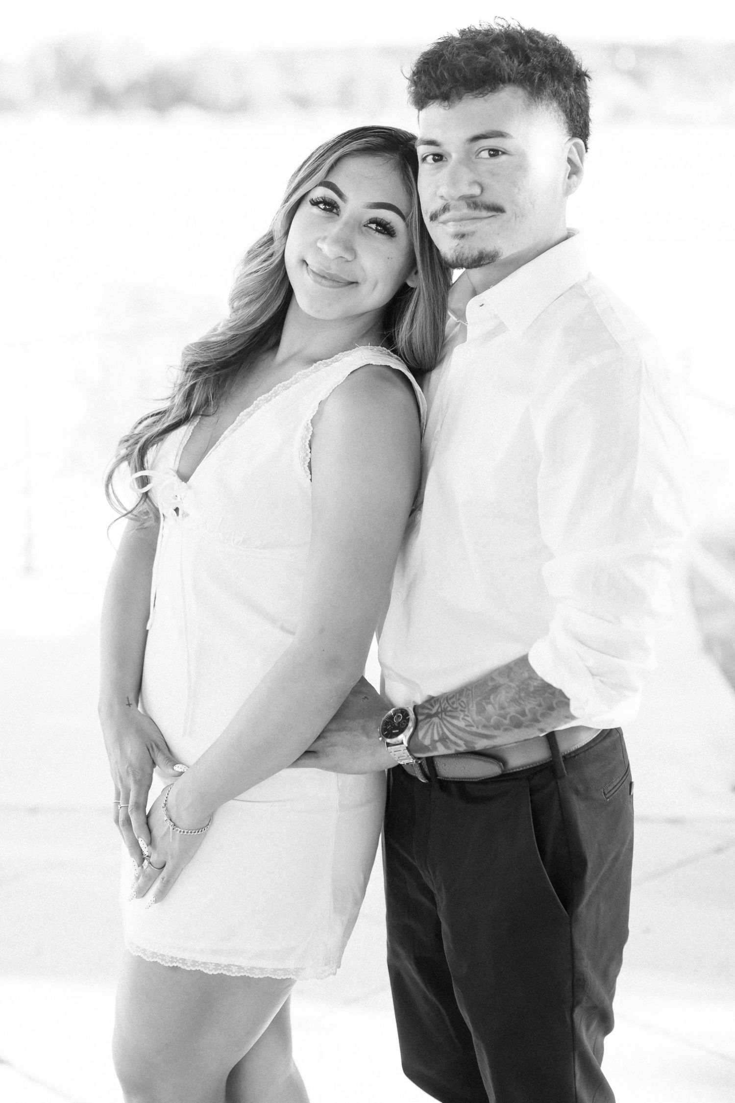 Black and white portrait of a young couple posing together, woman in white dress, man in white shirt and dark pants.