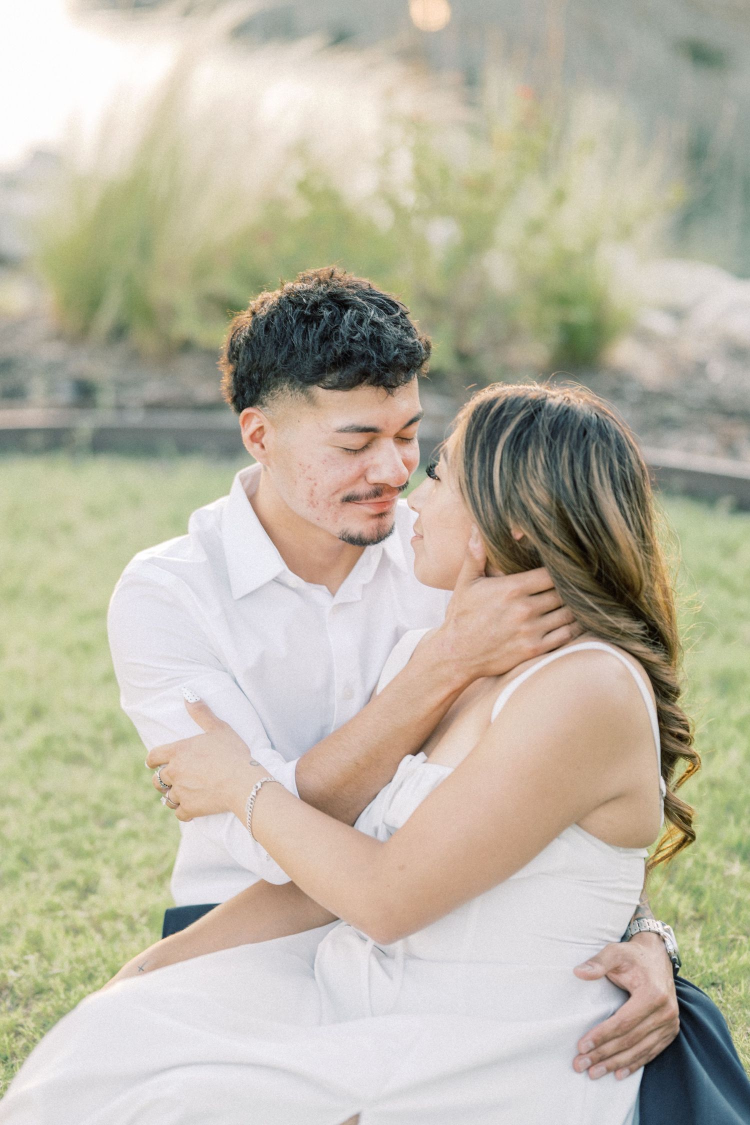 Couple sharing a tender almost-kiss moment while seated on grass during a golden hour engagement shoot.