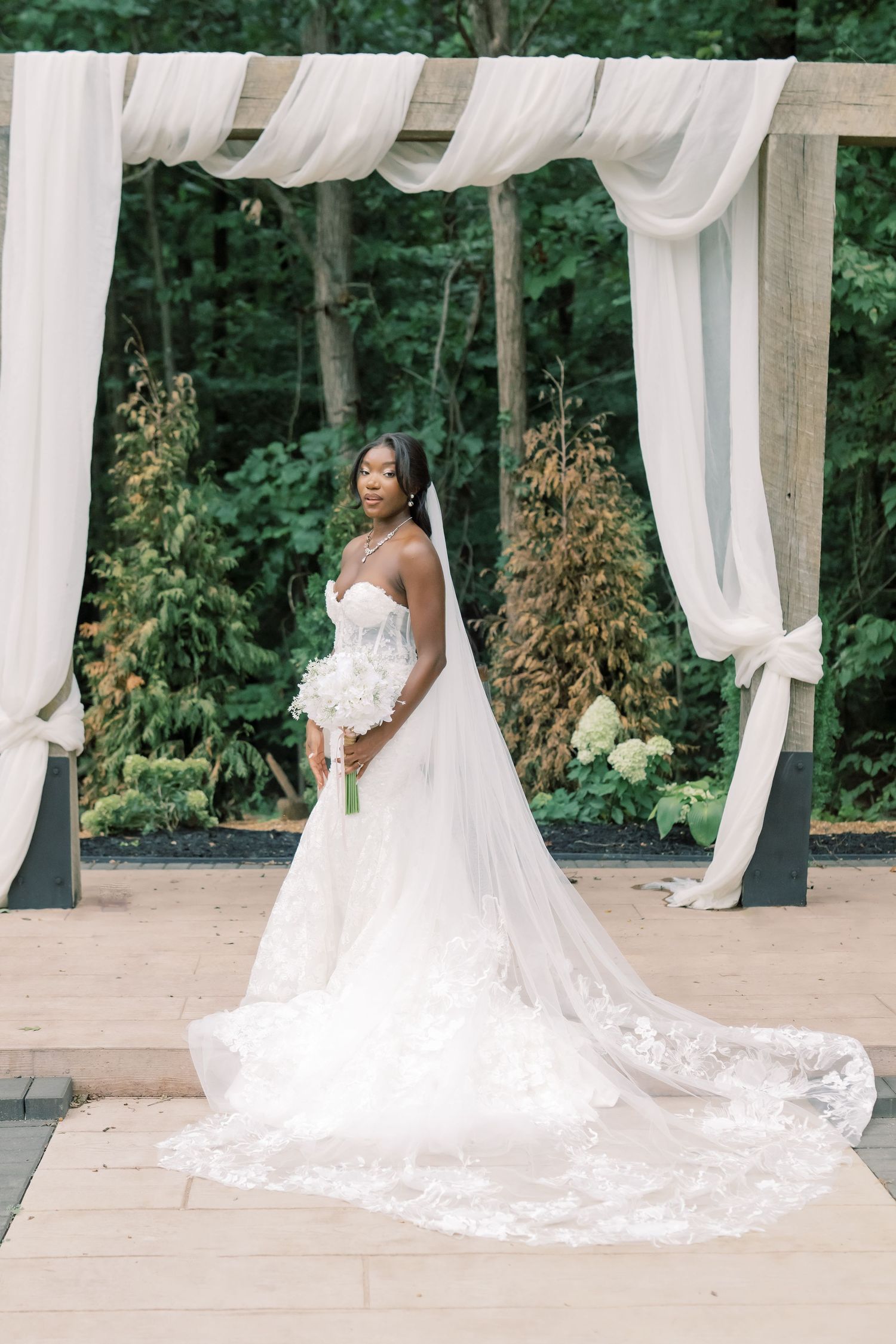 Elegant Black bride standing under white draped wooden arch, wearing strapless lace gown with cathedral veil and bouquet.