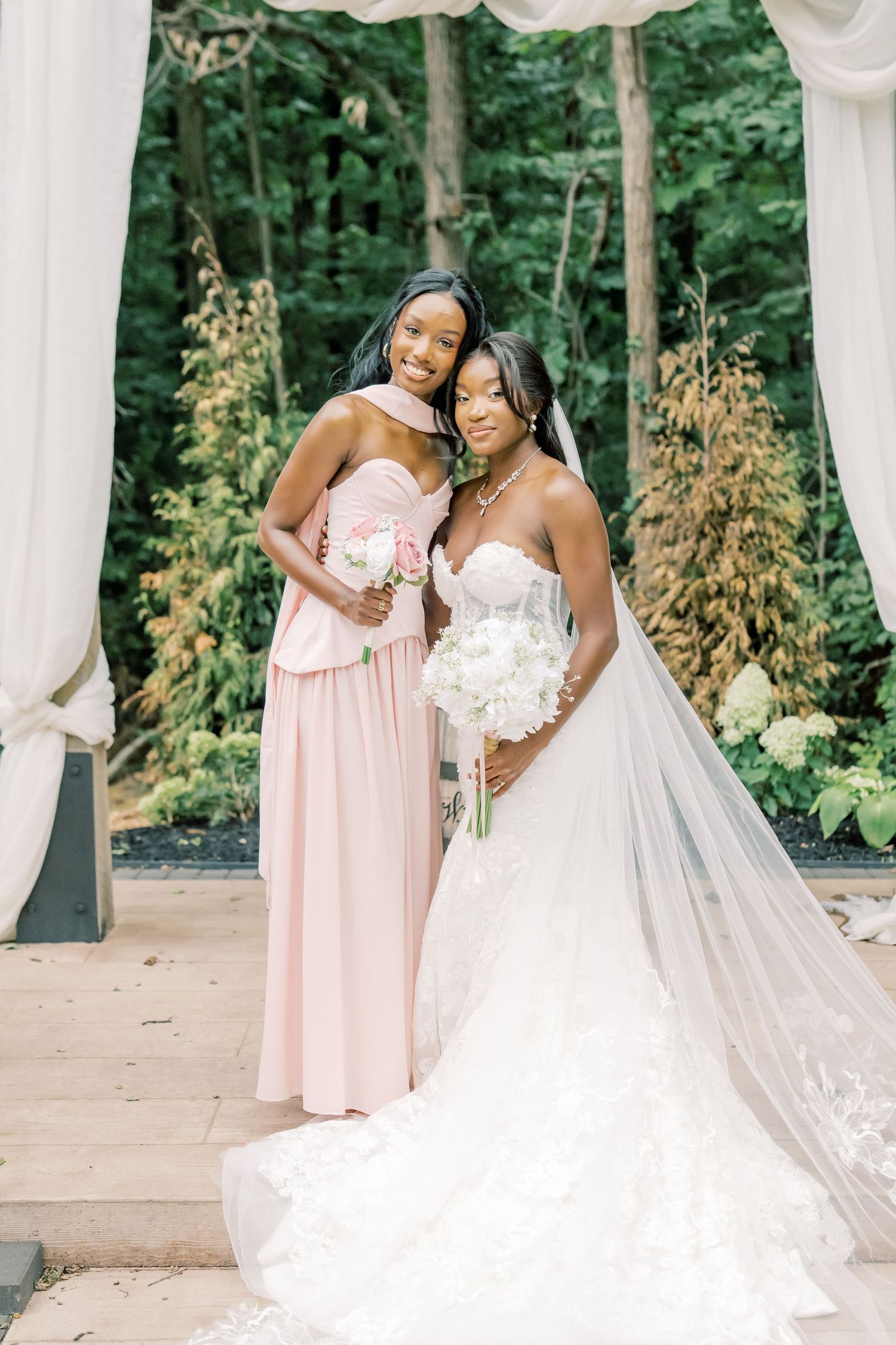 Bride in lace gown with veil and bridesmaid in blush pink dress pose together at outdoor wedding ceremony.