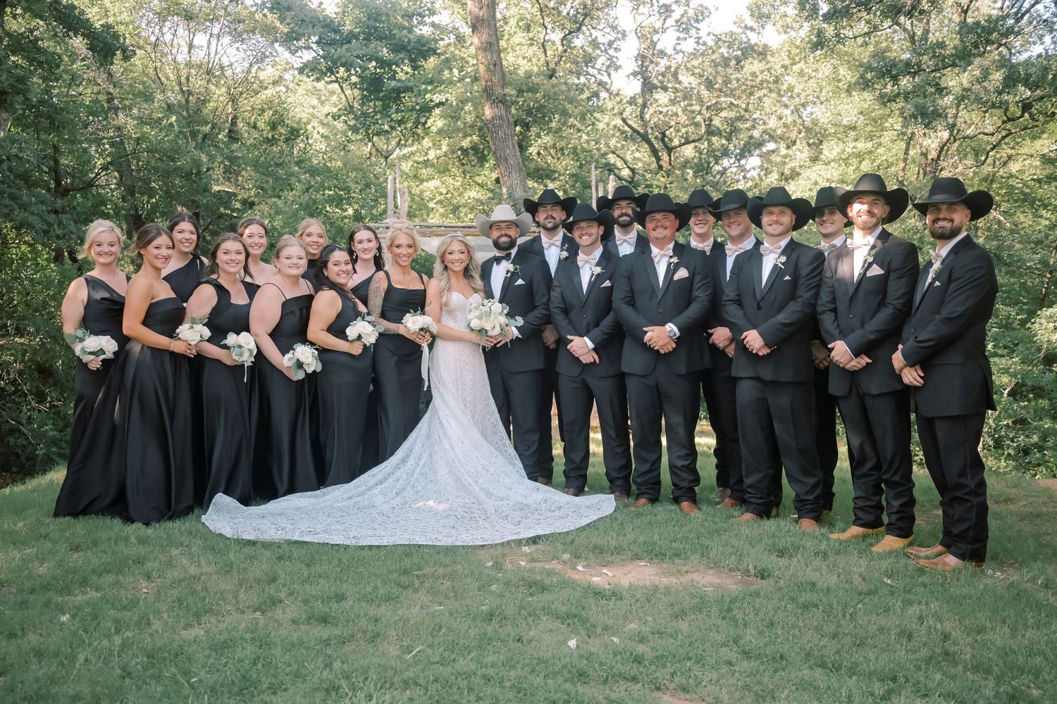Large wedding party in black attire with cowboy hats poses outdoors with bride in lace gown.